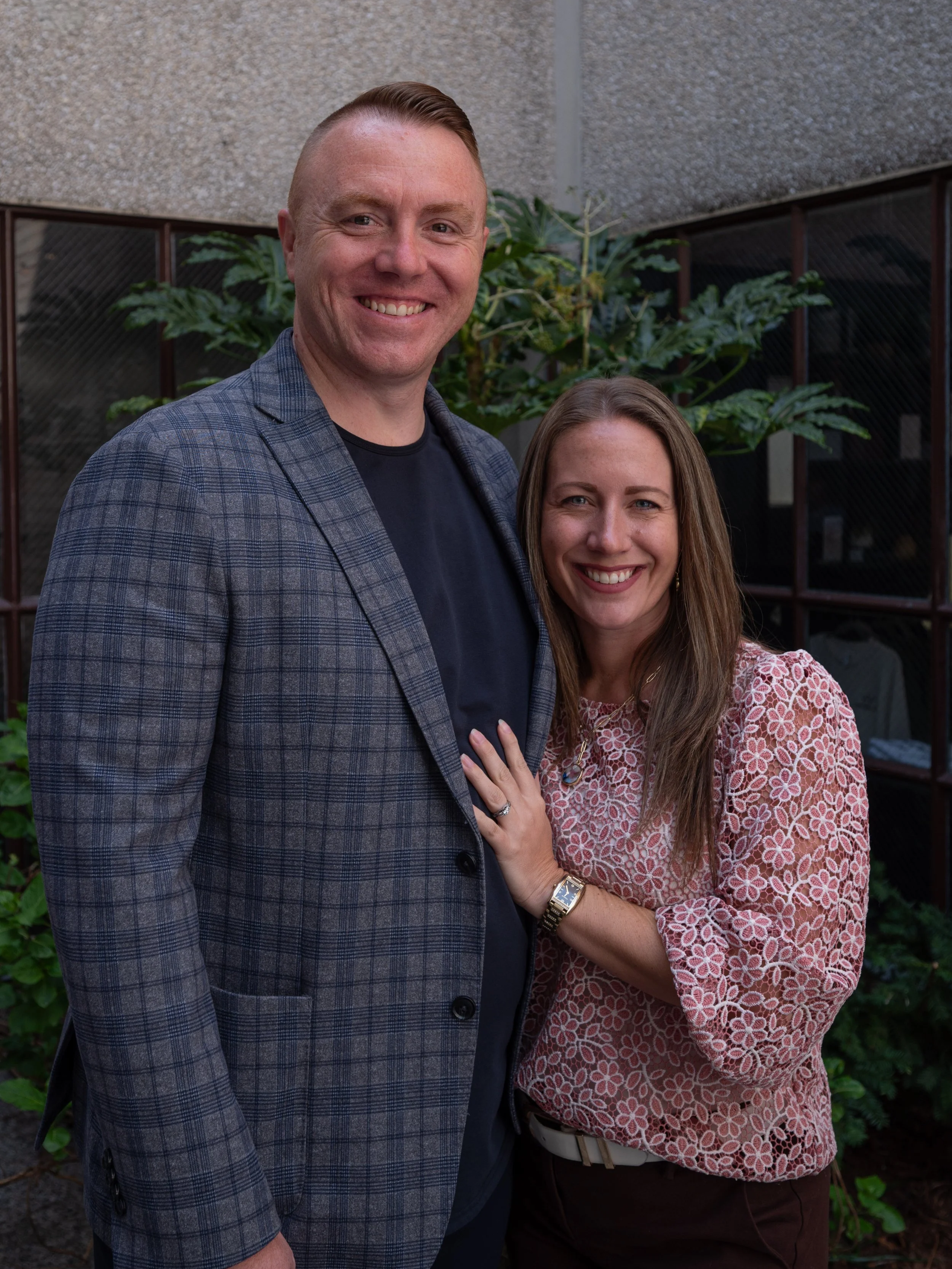 A smiling man in a checked blazer and a woman in a floral top standing close together outdoors, with green plants and a building wall in the background.