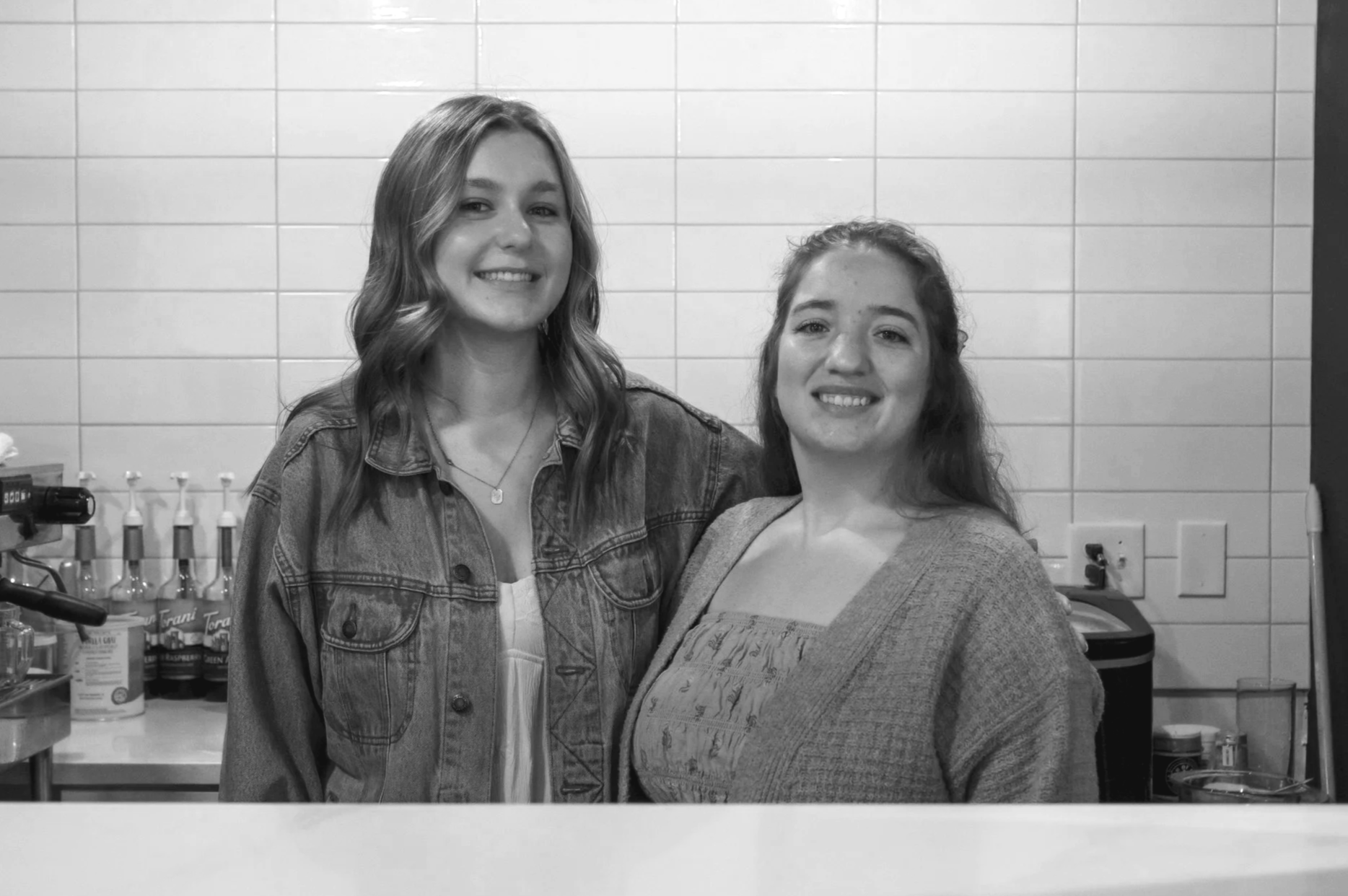 Two young women standing in a room with tiled walls, smiling at the camera. One has wavy hair and is wearing a denim jacket, the other has straight hair and is wearing a patterned top under a cardigan. Various bottles and objects are visible on the counter behind them.