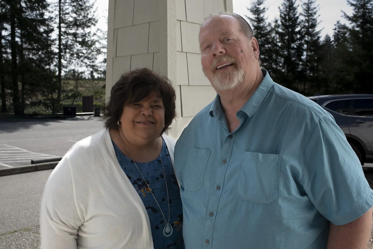 A woman and a man standing outside near a white building. The woman is wearing a white cardigan over a blue patterned shirt and a necklace, and the man is wearing a light blue shirt. There are trees and parked cars in the background.