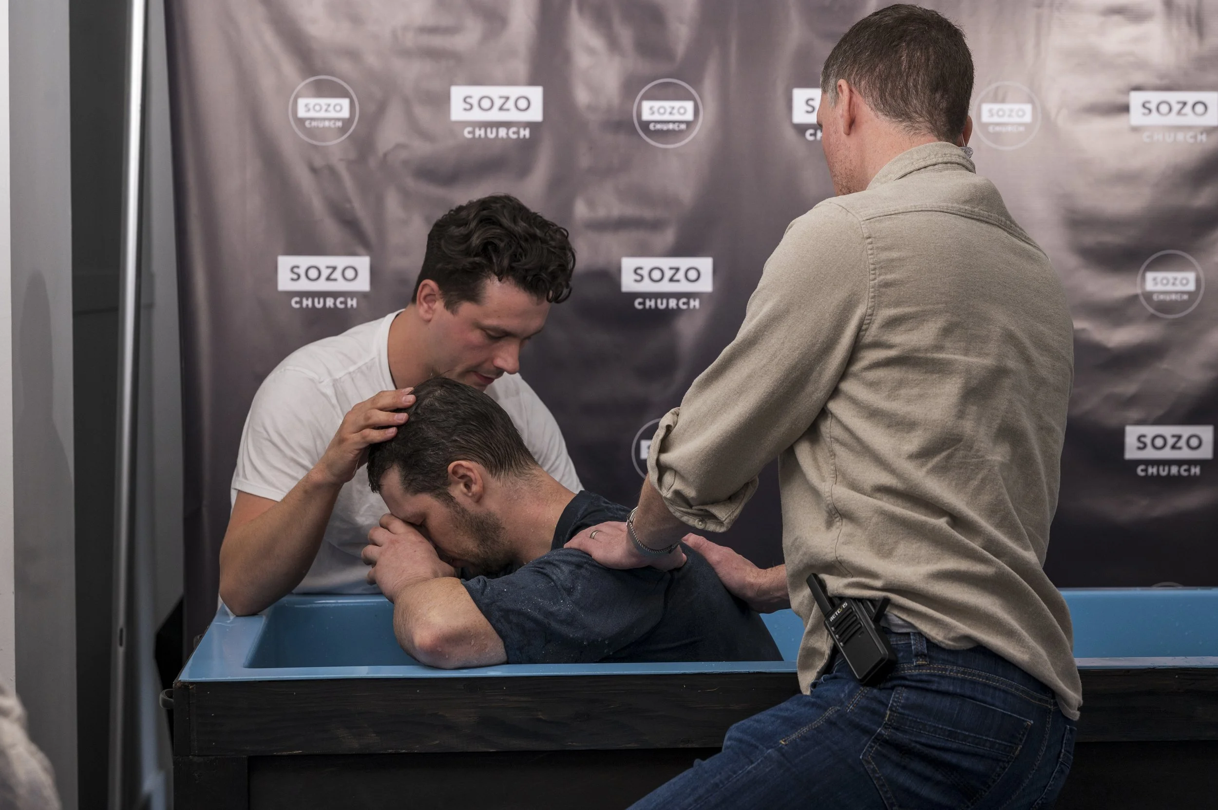 A man is being baptized in a church, with others present around him, as he kneels at a baptismal font and leans forward with his eyes closed.