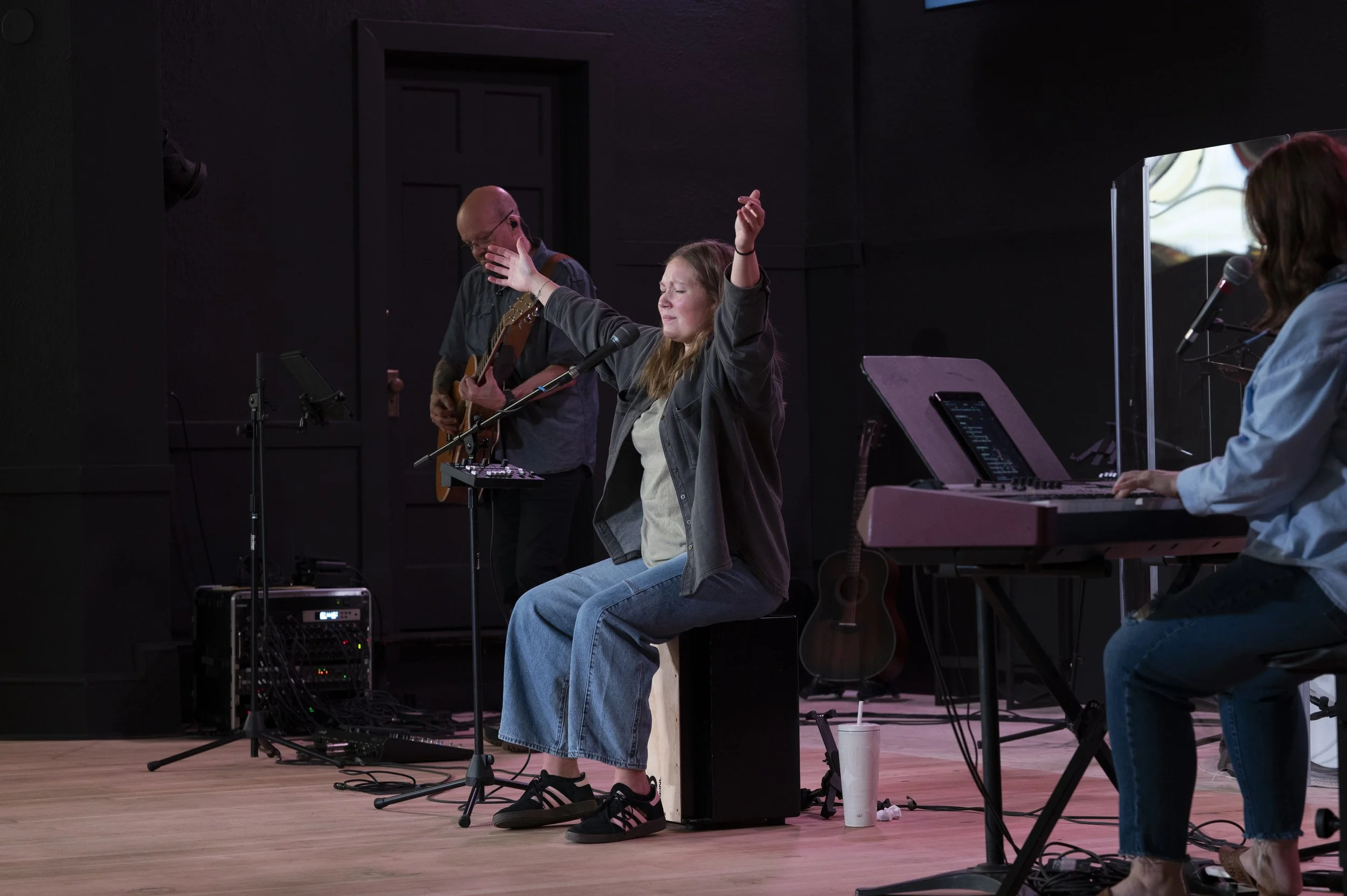 A woman is sitting on a stool with her eyes closed and arms raised as a man standing next to her plays an acoustic guitar and sings into a microphone. Other musicians are visible in the background on stage.