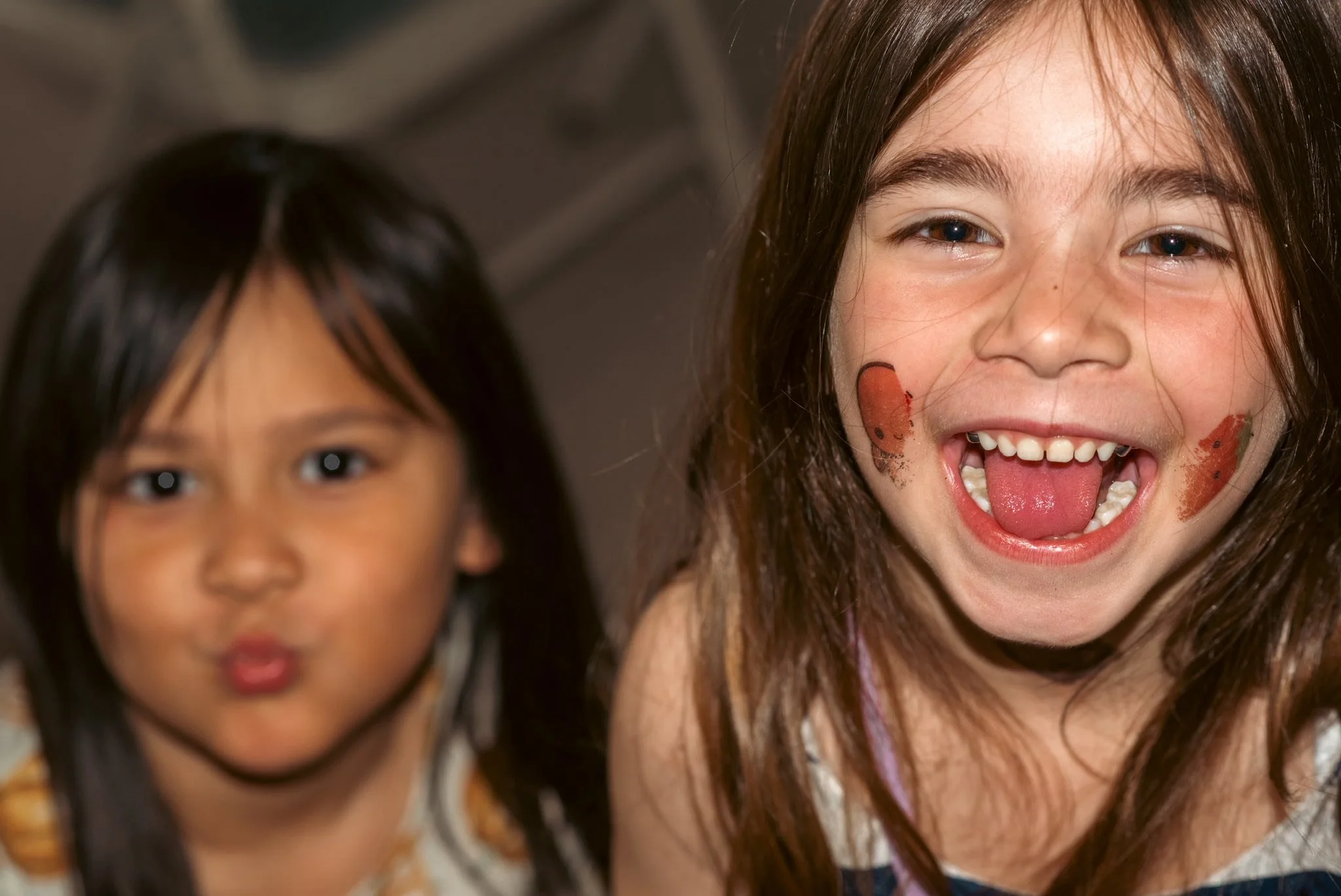 Close-up of two young girls, one smiling with her tongue out, the other slightly blurred, both with long dark hair.