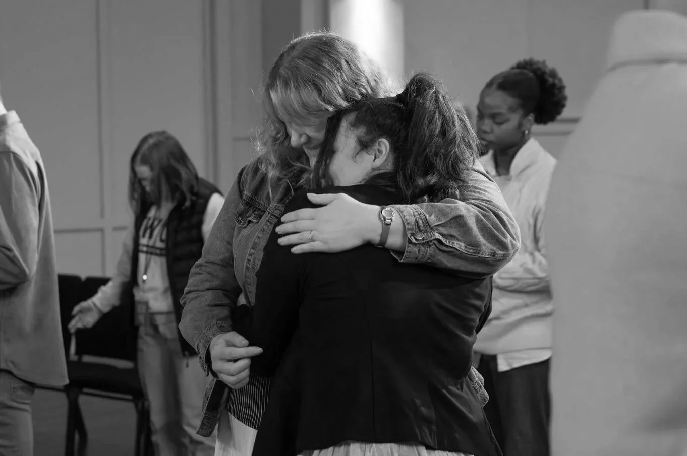 Two women hugging each other in an emotional embrace in a room with other people in the background.