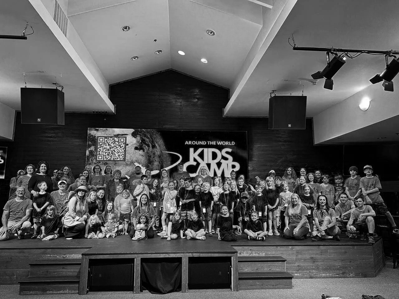 Group photo of children and adults on stage at a Kids Camp event, with a large sign and QR code behind them reading "Around the World Kids Camp."