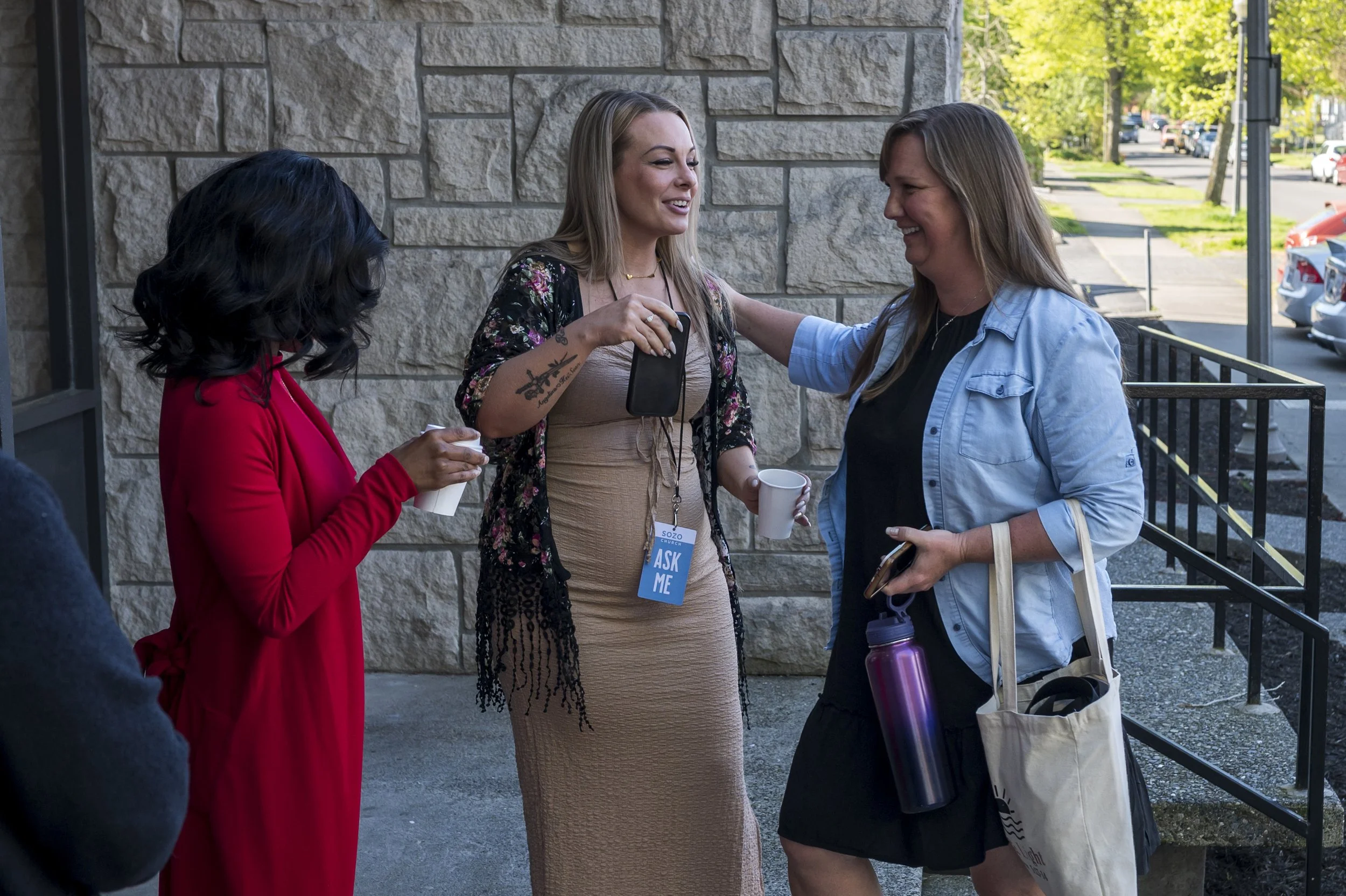 Three women are standing outdoors, engaged in conversation. One woman is wearing a red blazer, another is in a beige dress with a floral black cover-up, and the third woman is in a light blue shirt with a black inside shirt. They are smiling and holding cups, with trees and parked cars in the background.