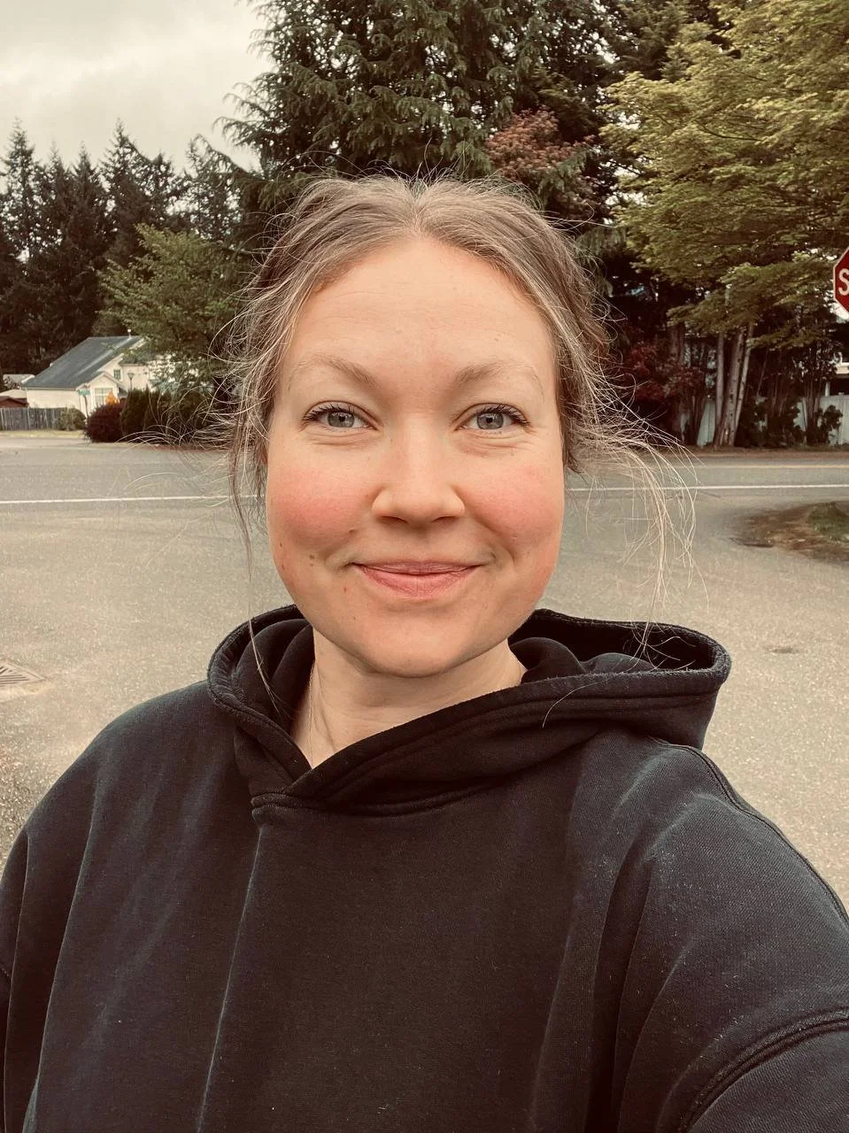 A woman with a slight smile, wearing a black hoodie, standing outdoors with trees and houses in the background.