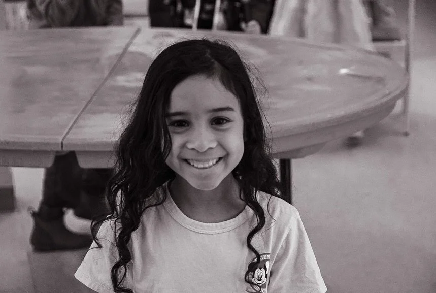 A young girl with long curly hair smiling at the camera in an indoor setting with a round wooden table in the background.