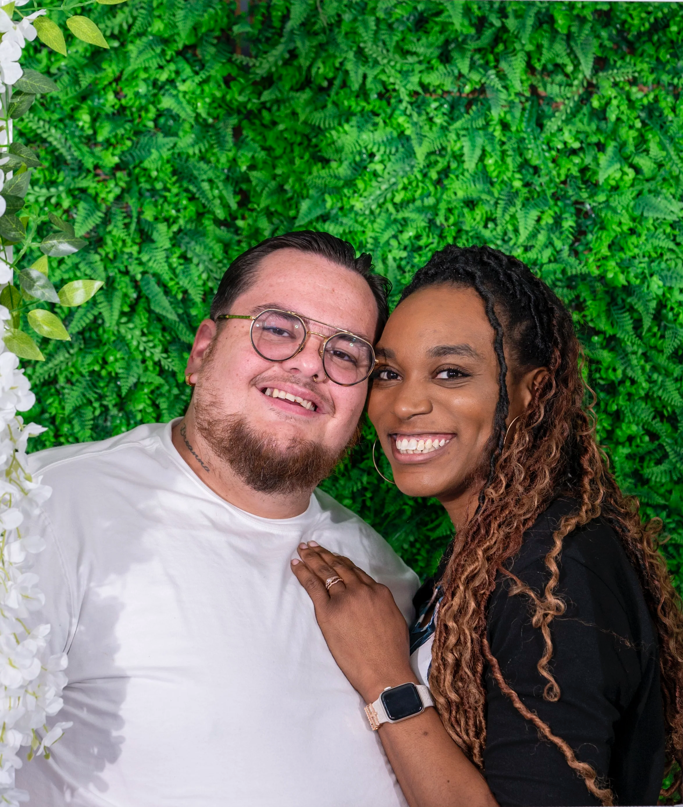 A man and woman smiling close together with a green fern wall background, partially visible, as they pose for a selfie. The man has glasses, light skin, and facial hair; the woman has darker skin and curly hair.