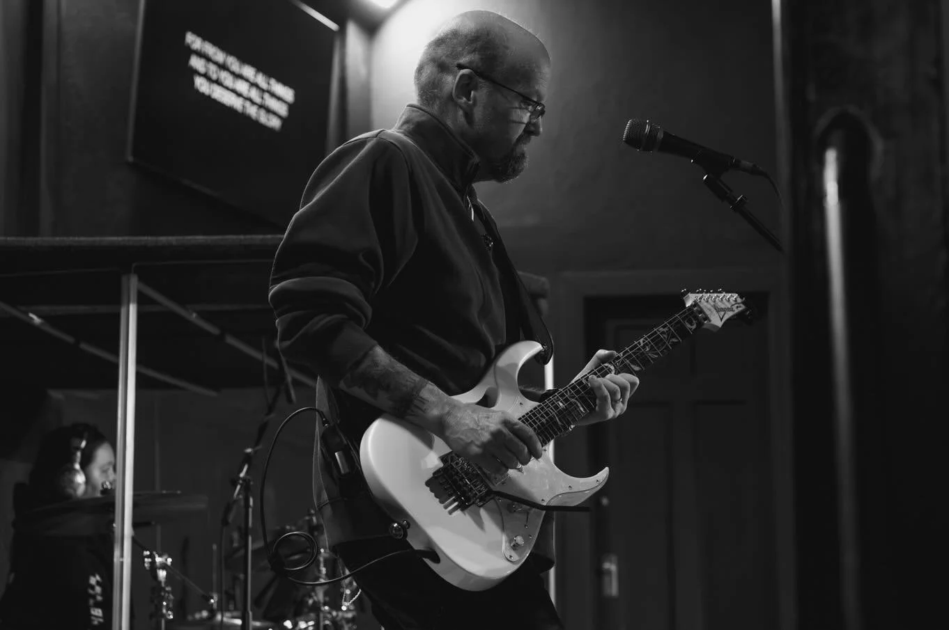 A man with glasses and a beard playing an electric guitar in a dimly lit room, with a woman in the background near a microphone and a monitor screen. The scene is in black and white.