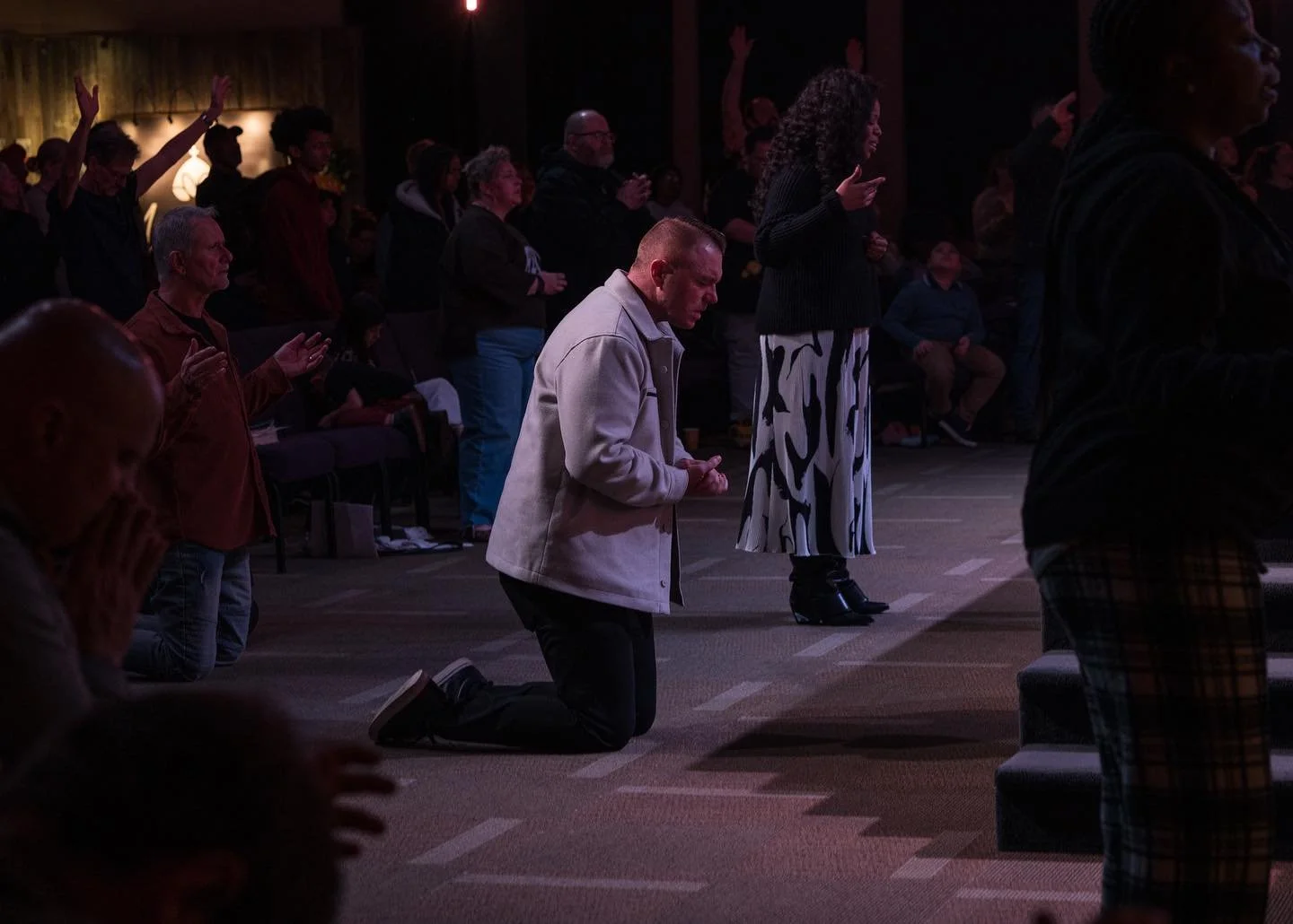 People kneeling and praying in a dimly lit indoor space, some with eyes closed, hands clasped, and others with hands raised, during a religious or spiritual gathering.
