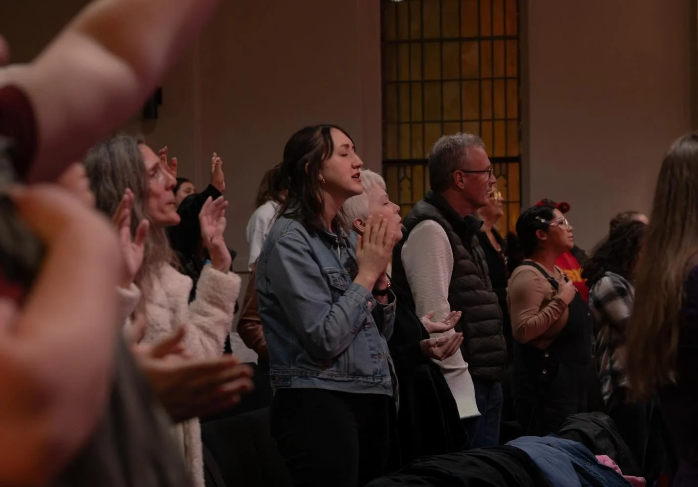 People standing in a church, engaged in prayer or worship, with some eyes closed, hands clasped or raised, and expressions of devotion.