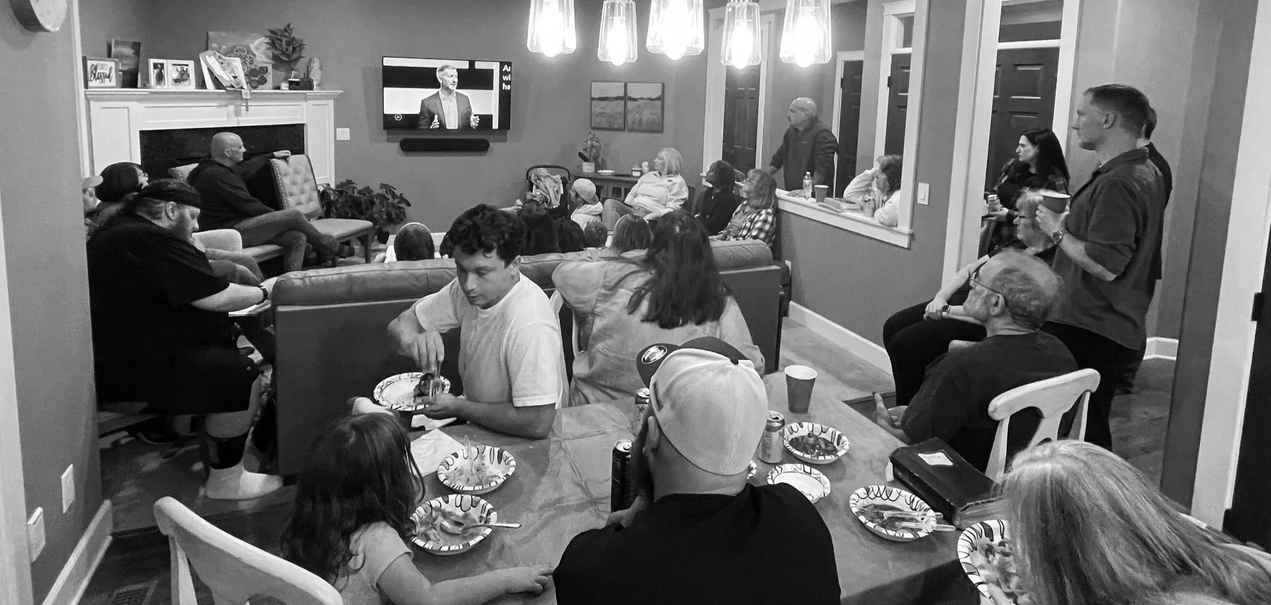 A group of people gathered in a living room watching a television, some sitting on chairs and couches, others standing, with a fireplace and decorated mantel in the background.