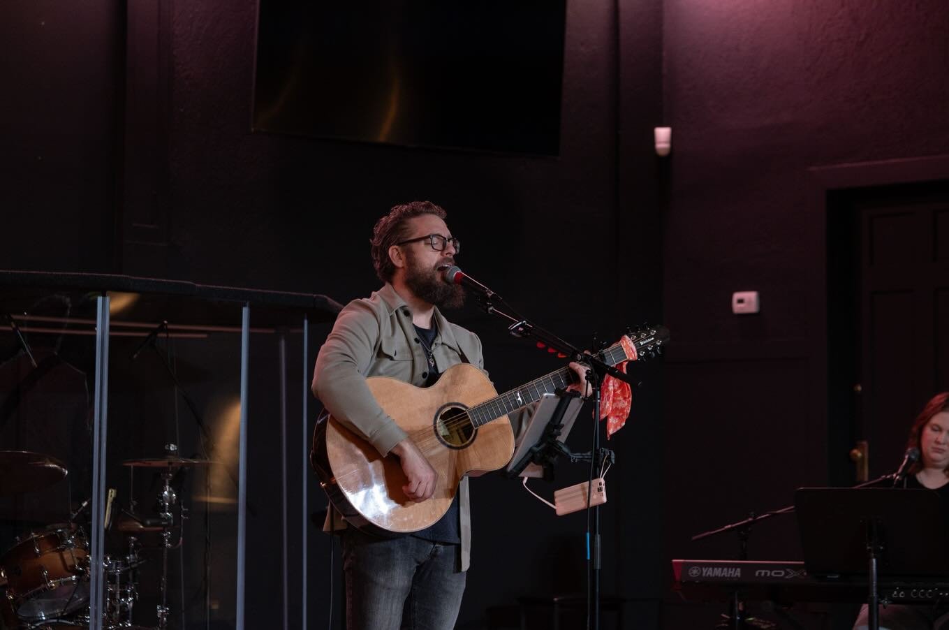 A man with glasses and a beard playing an acoustic guitar and singing into a microphone on stage, with music equipment and a woman playing a keyboard nearby.