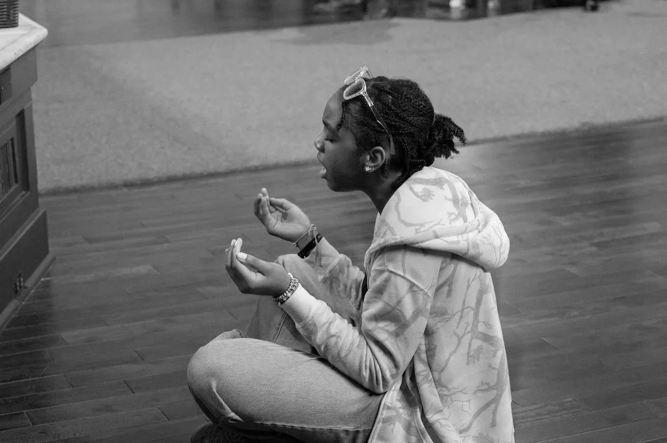 A young woman with glasses on her head sitting cross-legged on the wooden floor, with her eyes closed and hands raised in prayer or meditation.