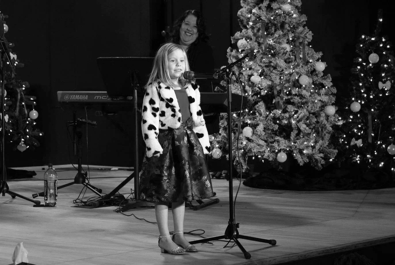 A young girl singing at a microphone during a Christmas performance, with Christmas trees decorated with ornaments in the background, and a keyboard performer smiling behind her.