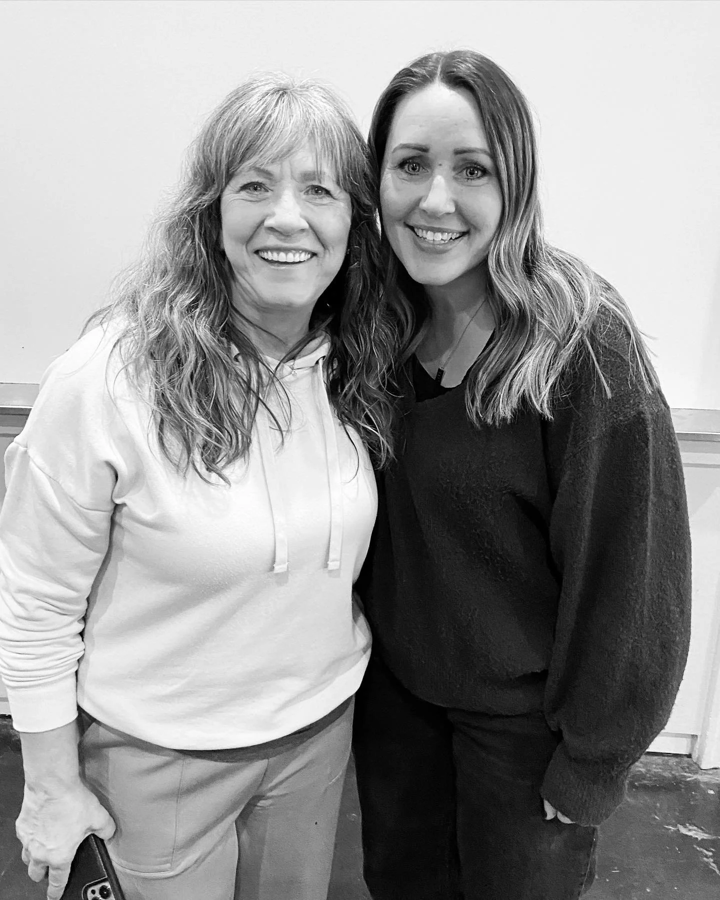 Two smiling women with long hair standing close together, one wearing a light hoodie and the other a dark sweater, against a plain wall.