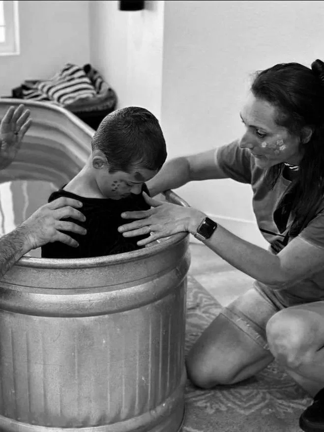 A young boy dressed in dark clothing is being baptized or immersed in a large bathtub, with a woman kneeling beside him and supporting him during the process.