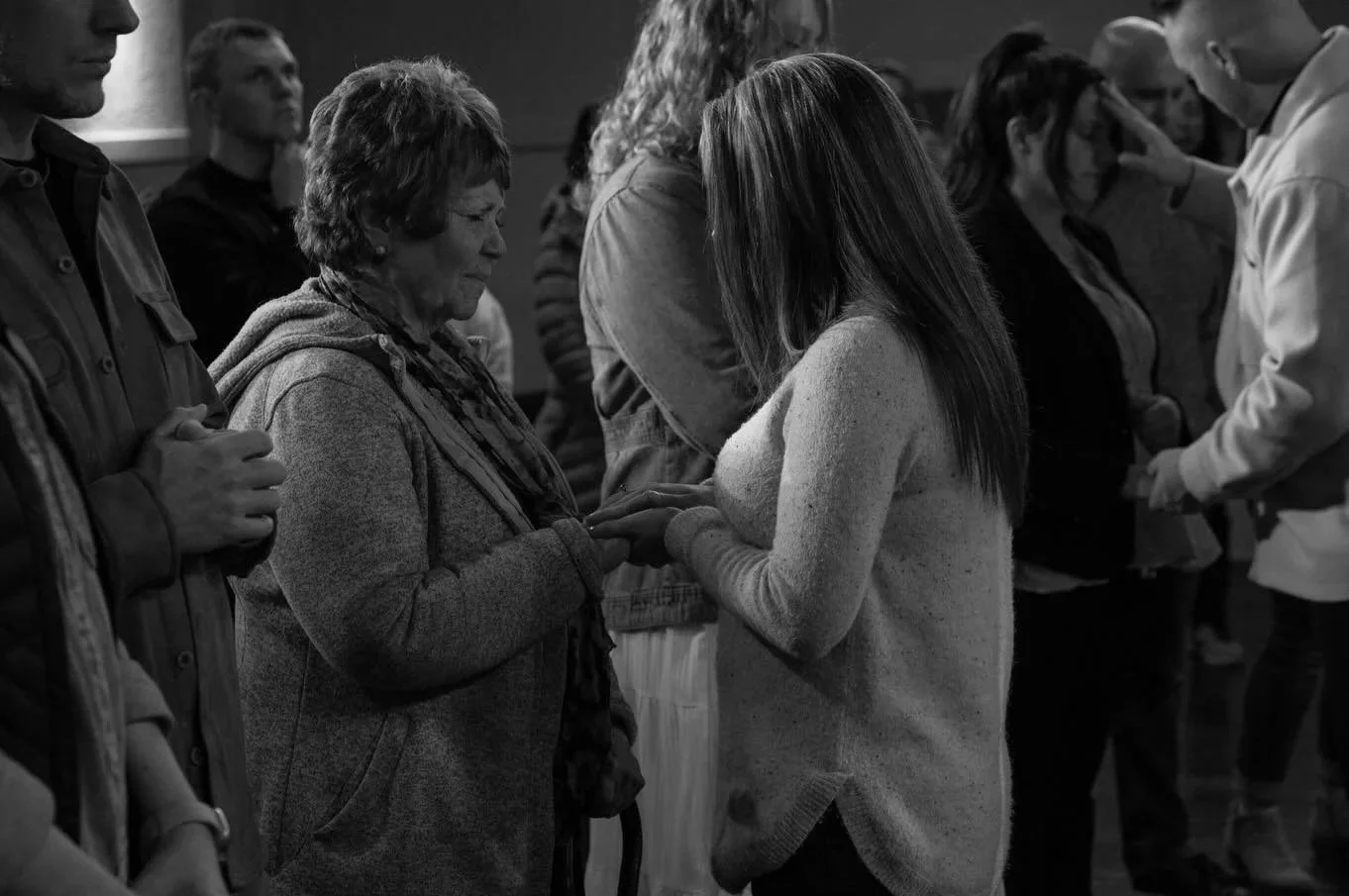 A group of people standing close together with heads bowed, engaging in a prayer or moment of reflection, with one woman holding another woman's hands in a comforting gesture.