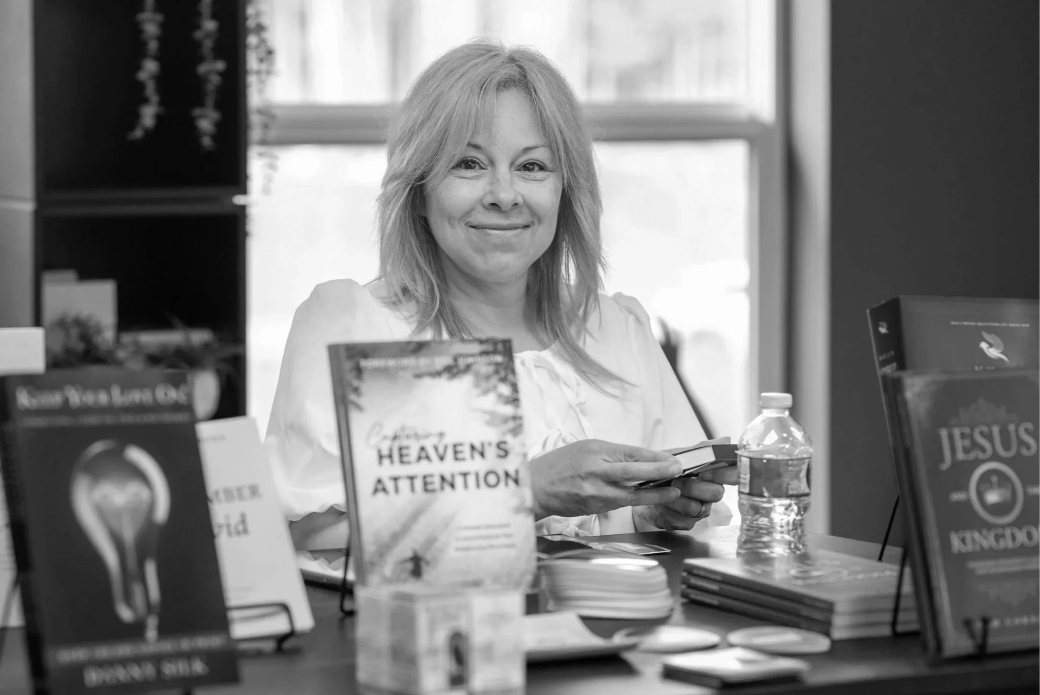 A woman with shoulder-length blonde hair sitting at a table surrounded by books, holding a book in her hands, with books and a water bottle in front of her in a room with large windows.