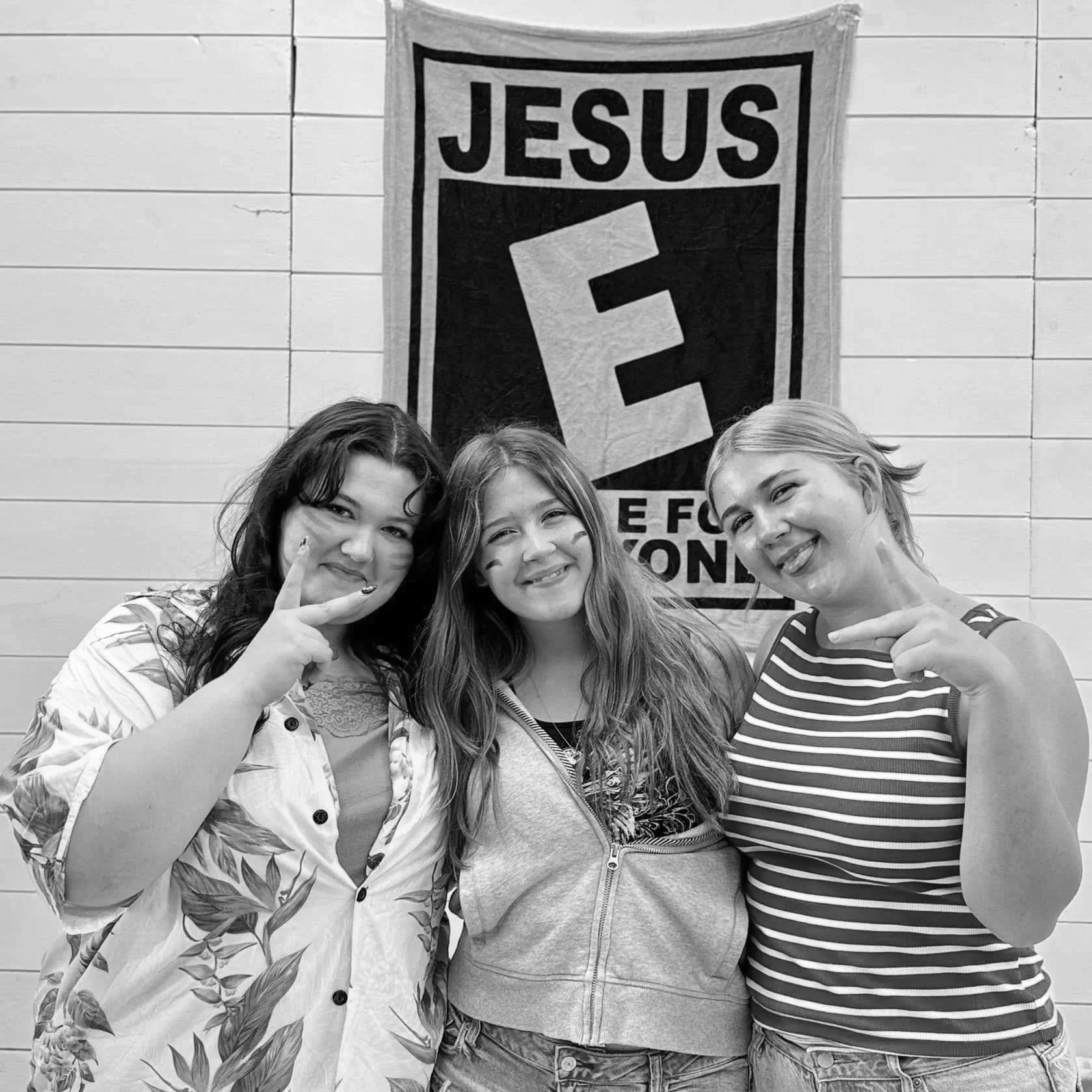Three young women smiling and posing together in front of a banner that reads 'JESUS' and has a large letter 'F' on it. All three women are pointing at themselves and appear happy.