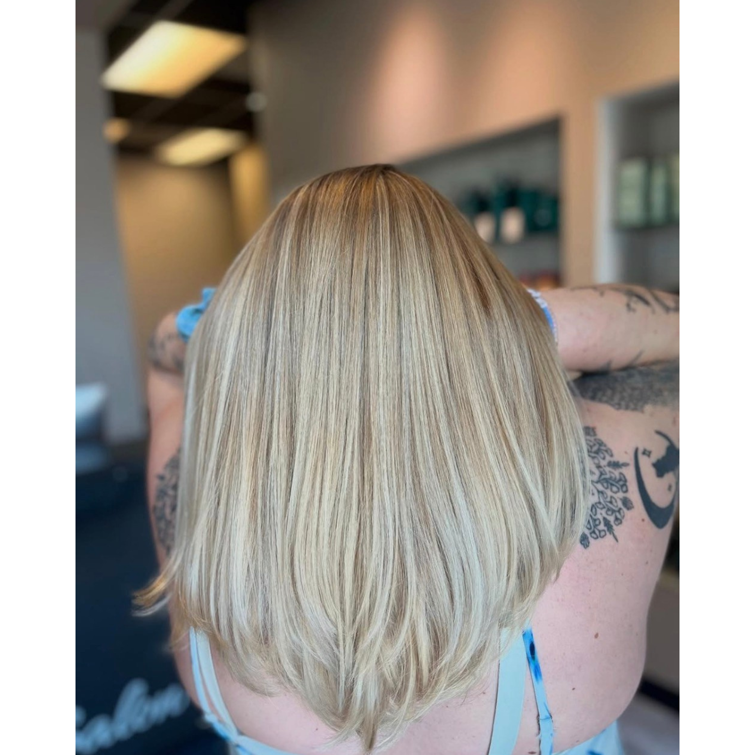 Blonde shoulder-length hair styled straight, with darker roots blending into lighter blonde ends, seen from the back in a hair salon.