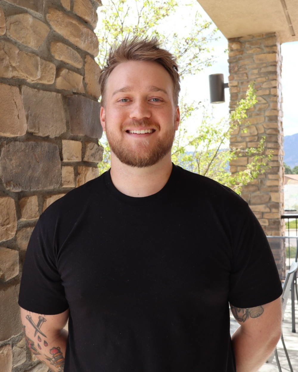 Close-up of a smiling young man with a beard, wearing a black t-shirt, standing outdoors near a stone wall with trees and mountains in the background.