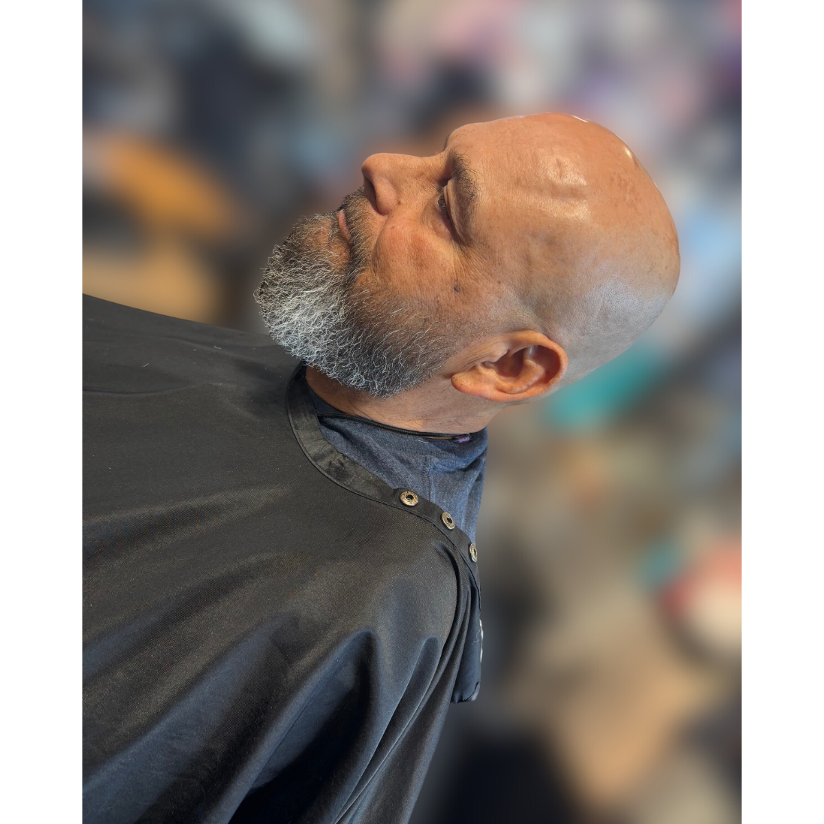 Side profile of a bald man with a gray beard, wearing a black barber cape in a barbershop.