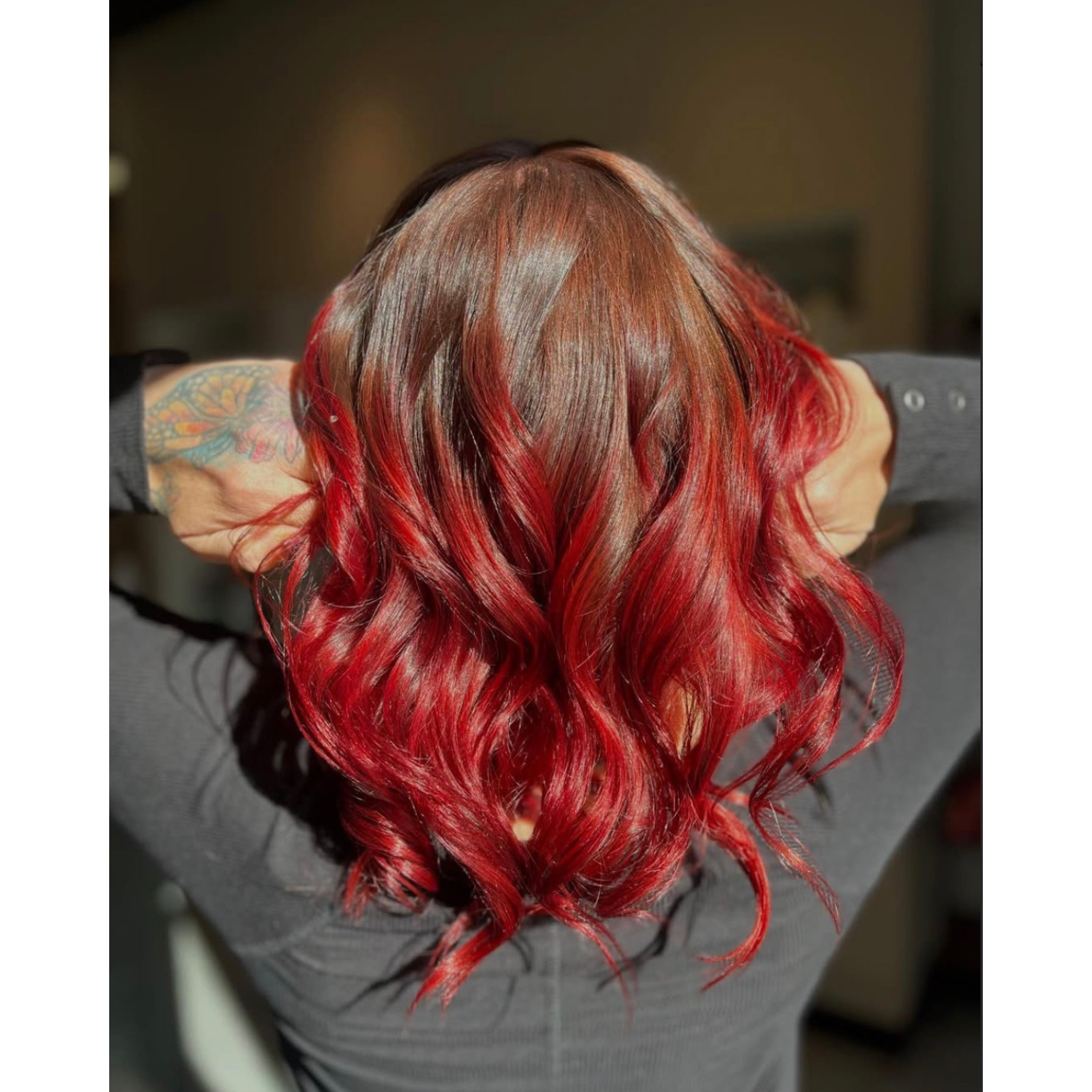 Back view of a woman with shoulder-length, wavy red hair, wearing a dark gray top, and showing a colorful tattoo on her left arm.