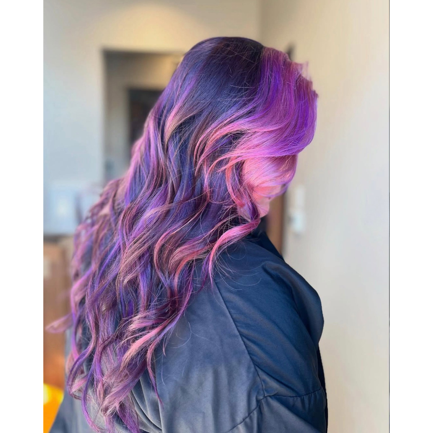 A woman with long, wavy hair dyed in shades of purple, pink, and blue, wearing a dark jacket.