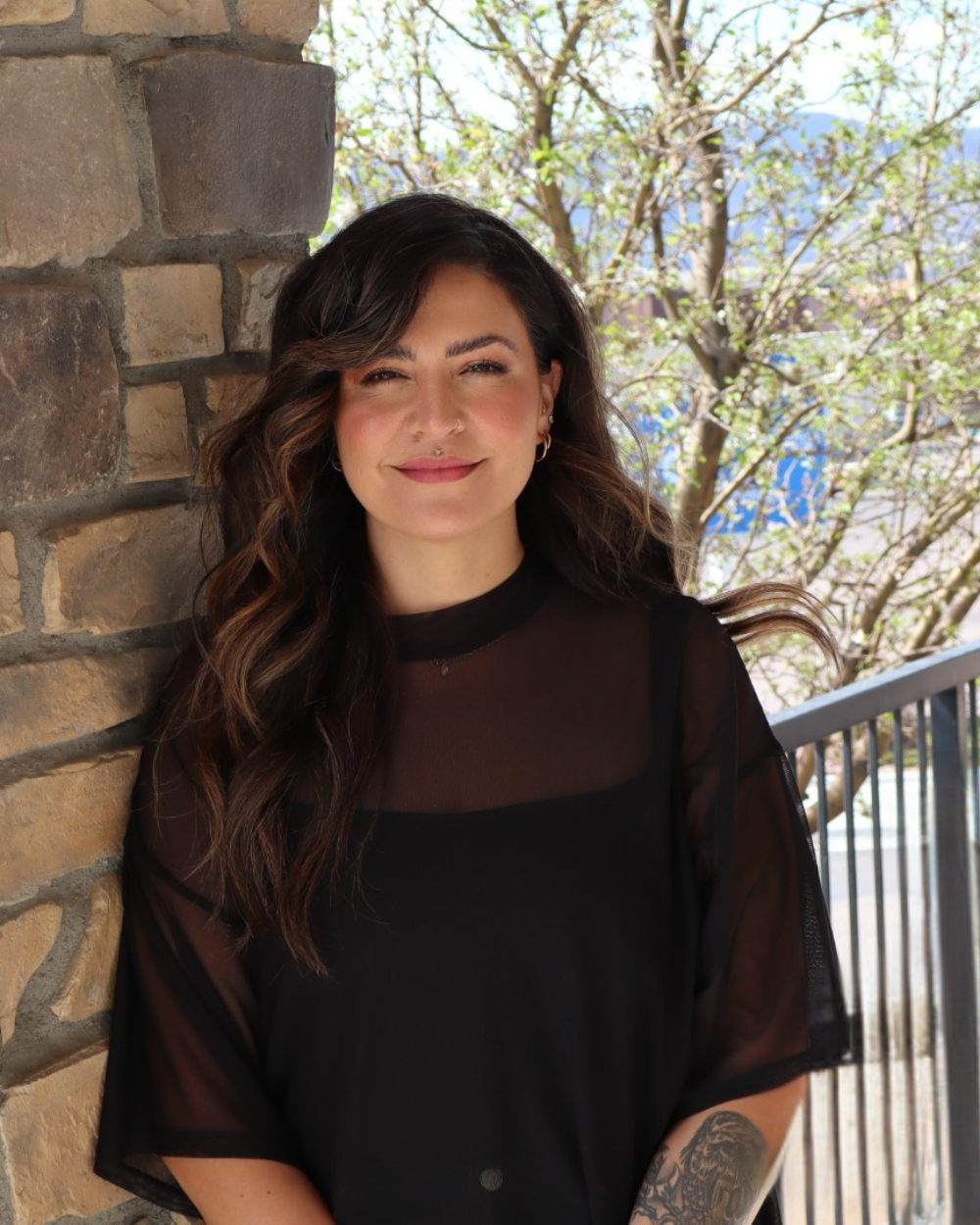 A woman with wavy brown hair and a nose piercing standing outdoors on a balcony with a stone wall to her left and greenery behind her.