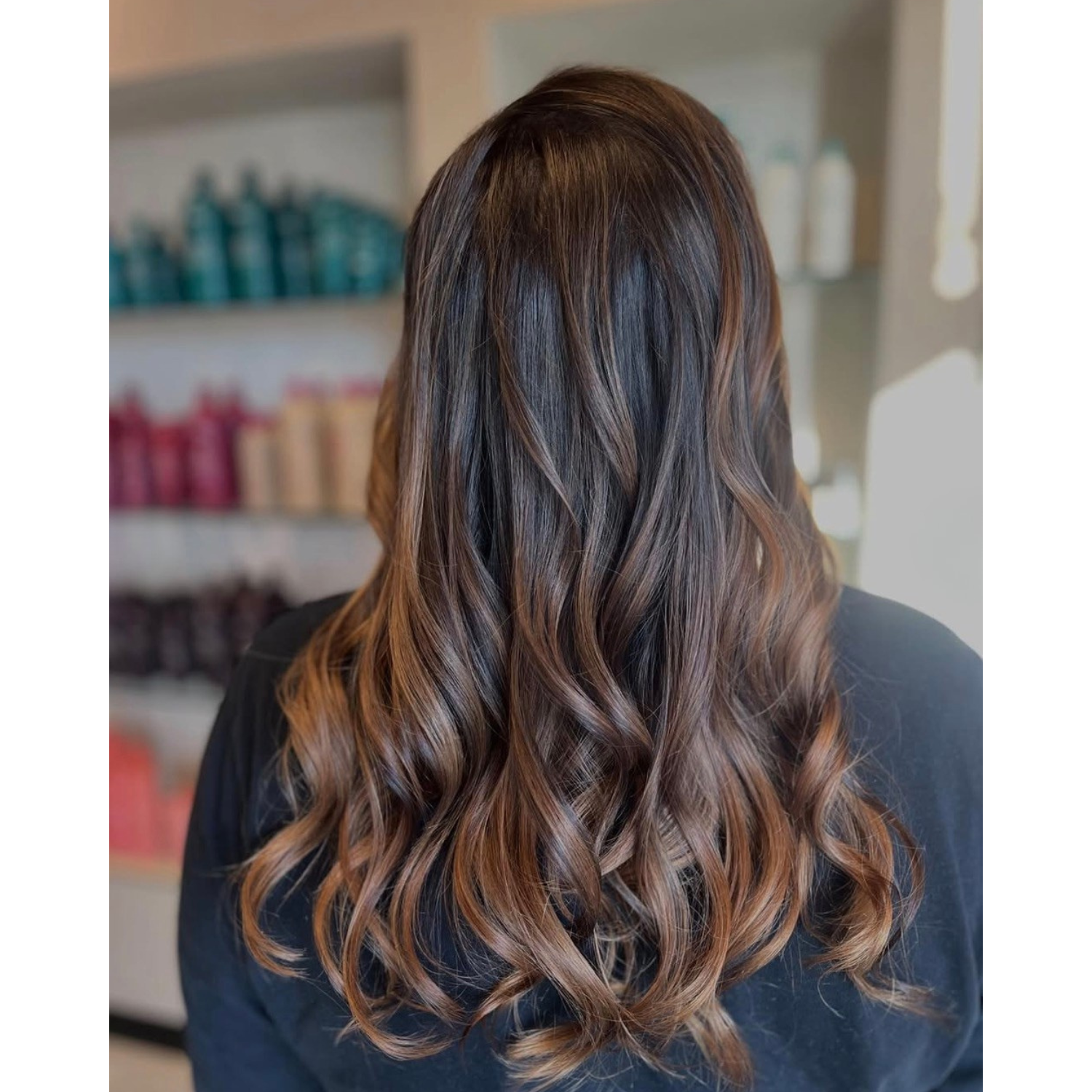 Back of a woman's head showing long wavy brown hair with caramel highlights, in a salon setting.