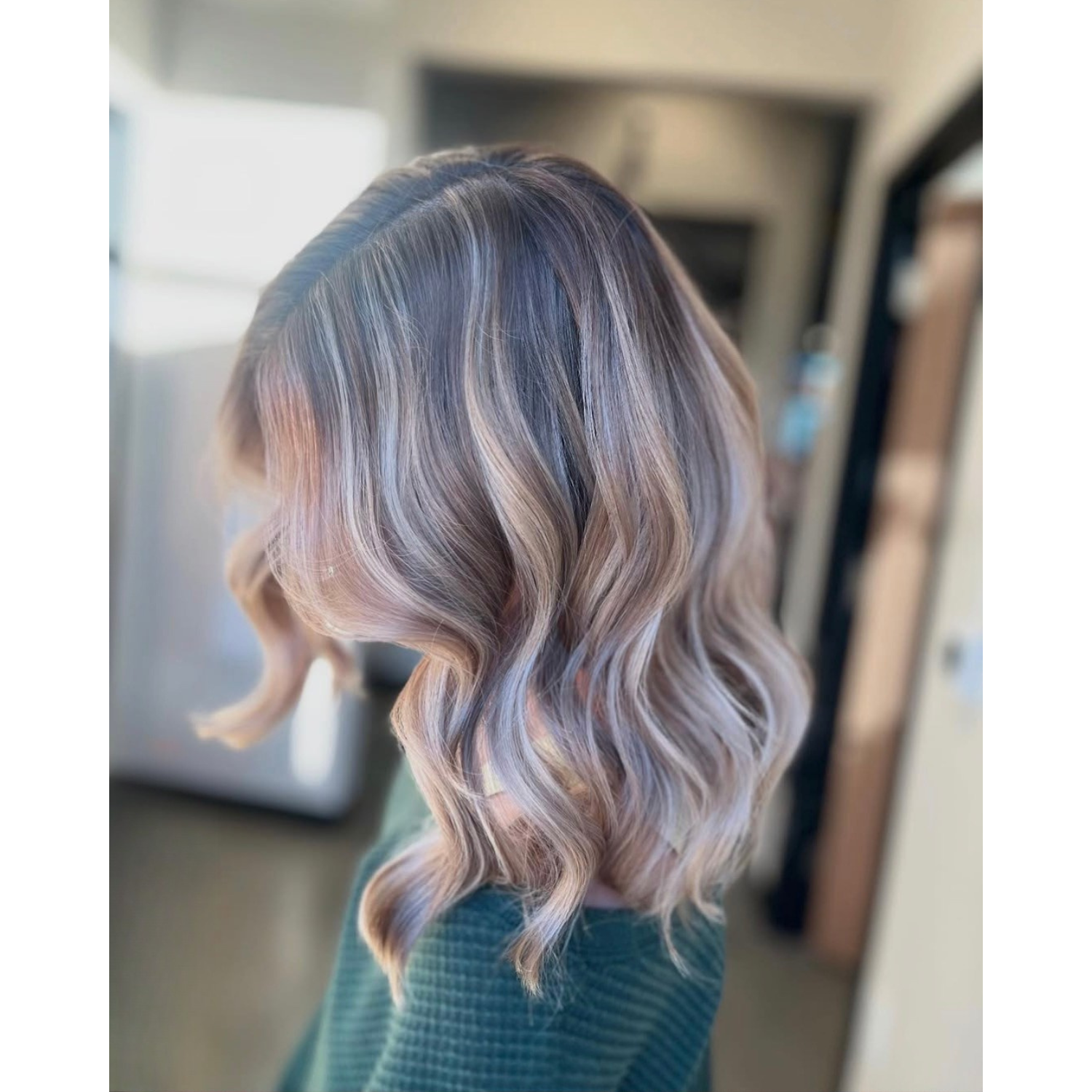 A woman with shoulder-length wavy hair styled in loose curls, featuring a blend of dark roots and light blond highlights.