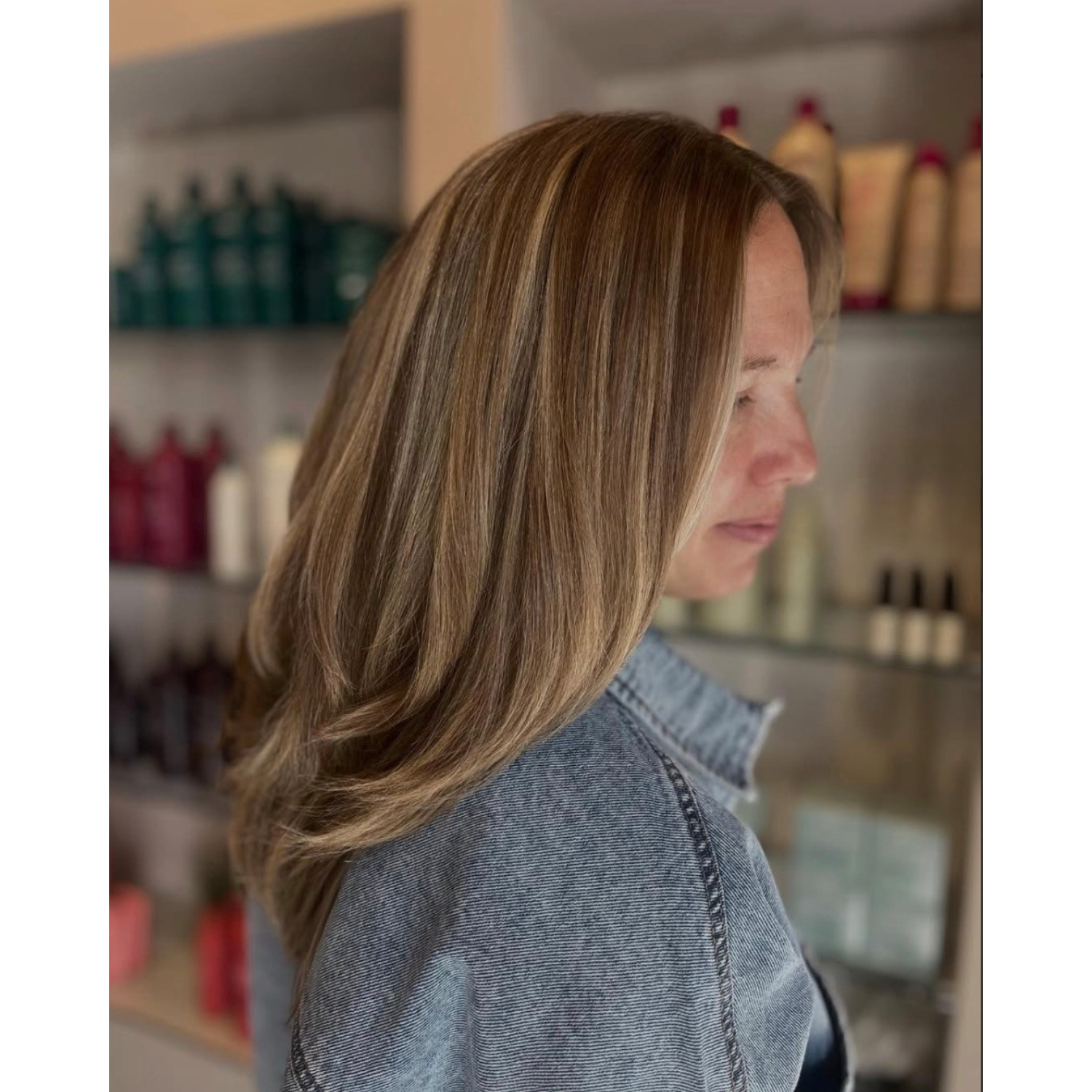 A woman with shoulder-length, straight, highlighted hair looking down, wearing a denim jacket, in what appears to be a hair salon.