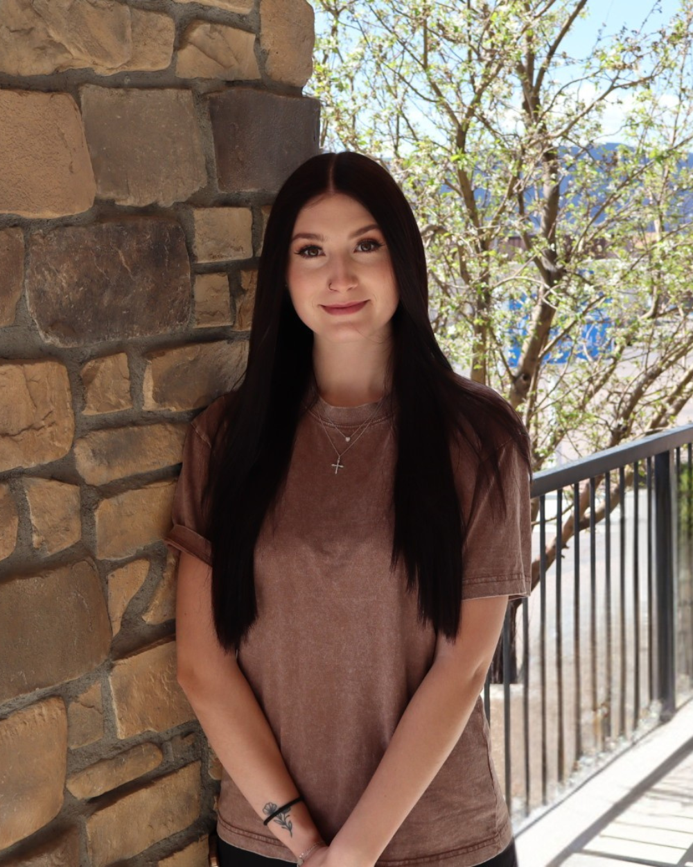 A young woman with long dark hair, wearing a brown T-shirt and silver necklaces, standing outdoors beside a stone wall, with trees and a black metal railing in the background.