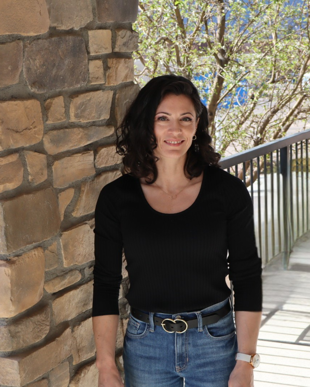 A woman with dark curly hair smiling, wearing a black top, jeans, a black belt with a heart-shaped buckle, and a watch, standing on a porch with a brick wall and a tree in the background.
