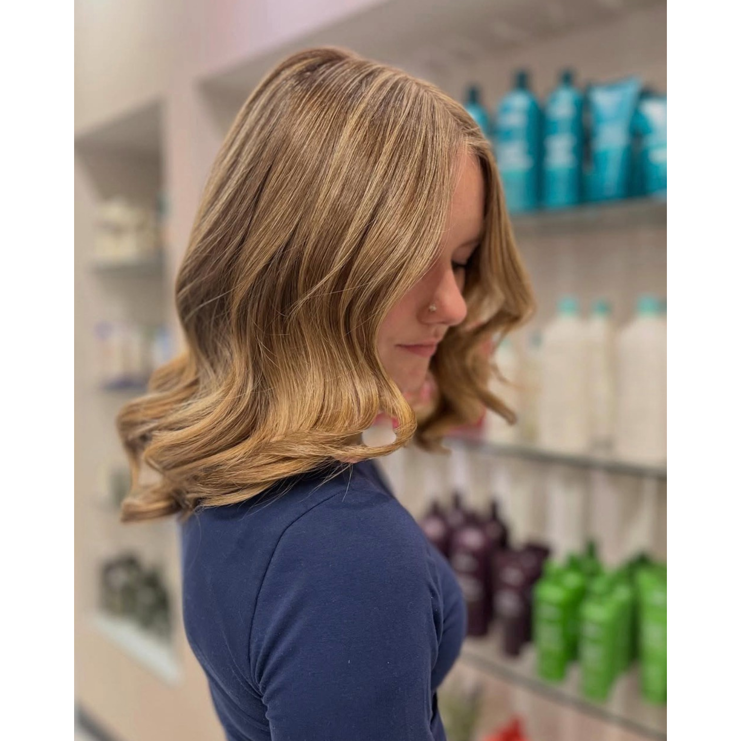 A woman with shoulder-length, wavy blonde hair, wearing a blue top, is standing in front of shelves stocked with bottles of hair care products. She has a nose piercing and is looking downward.