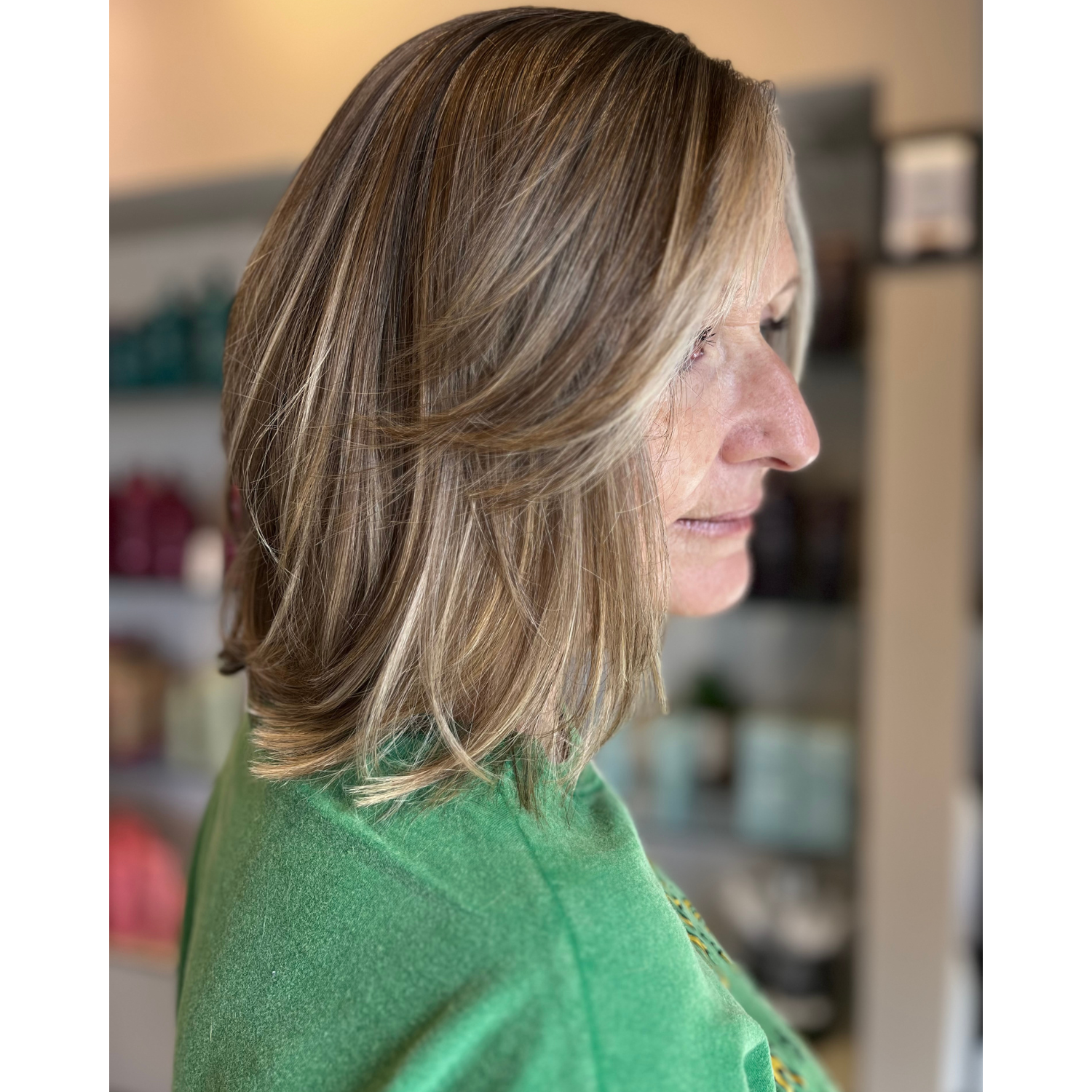Side profile of a woman with shoulder-length, layered brown hair wearing a green top, indoors with blurred background shelves.