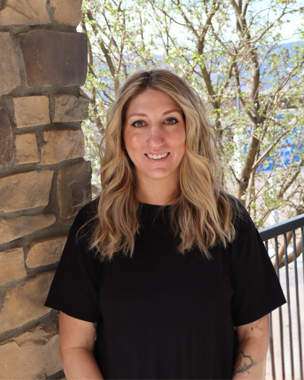 A woman with wavy blonde hair smiling outdoors on a balcony with trees and mountains in the background.