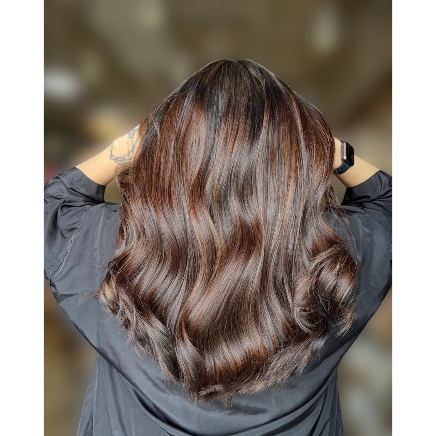 Back of a person with long, wavy, brown hair with subtle highlights, wearing a dark gray shirt and a smartwatch, standing against a blurred background.