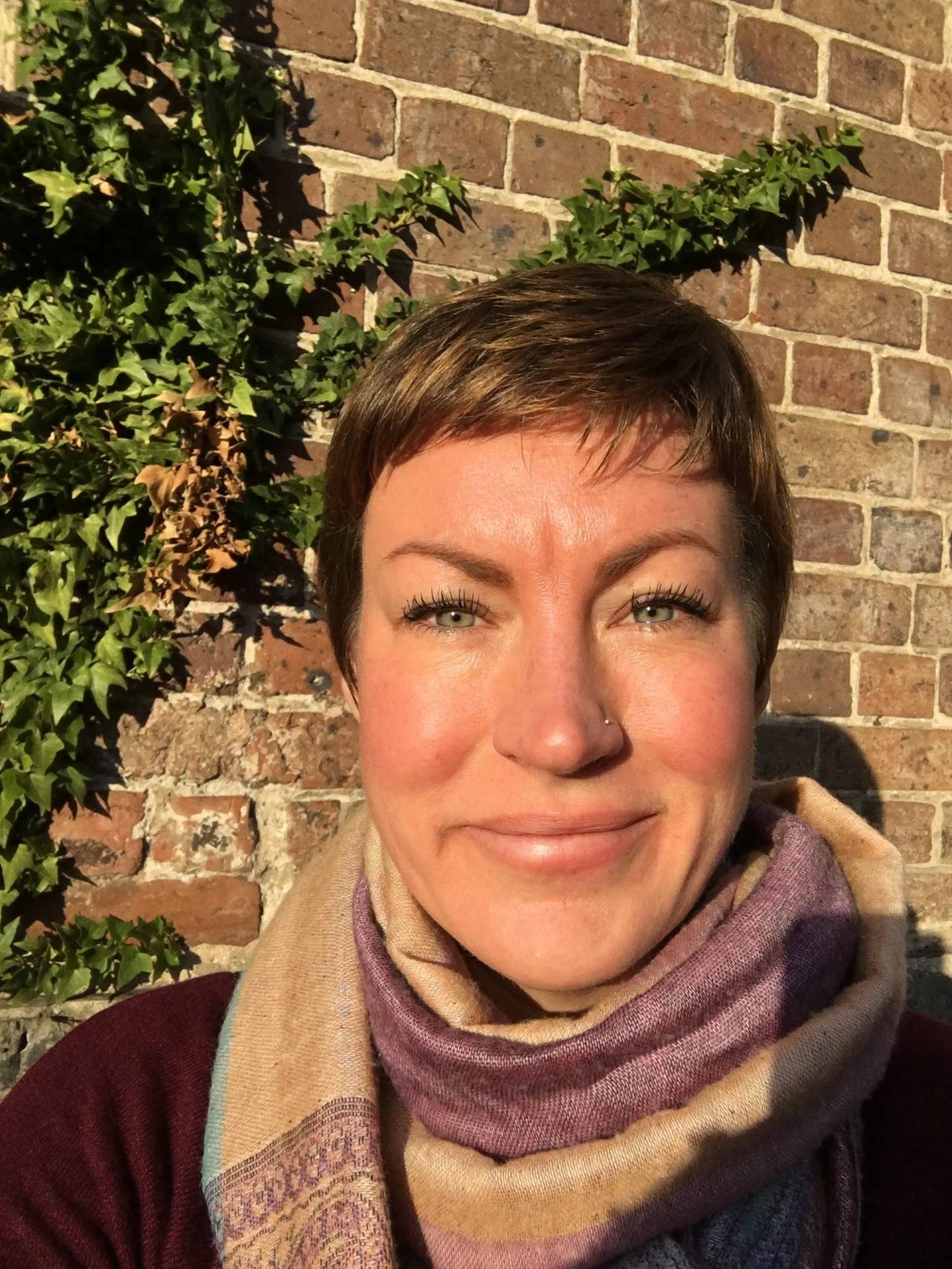 fair skinned female with short brown hair with a scarf, standing in front of a brick wall with ivy, smiling