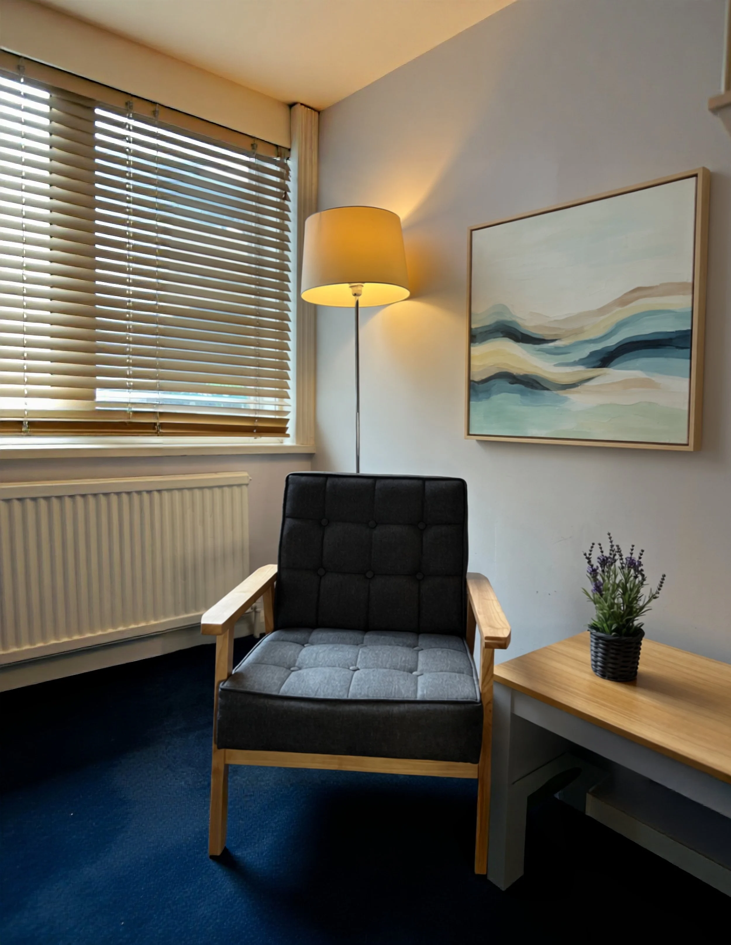 Mariana Aleksieva's therapy room with a black cushioned armchair, a wooden side table with a small potted lavender plant, a painting with abstract blue and beige waves on the wall, a floor lamp with a beige shade, and a window with wooden blinds.