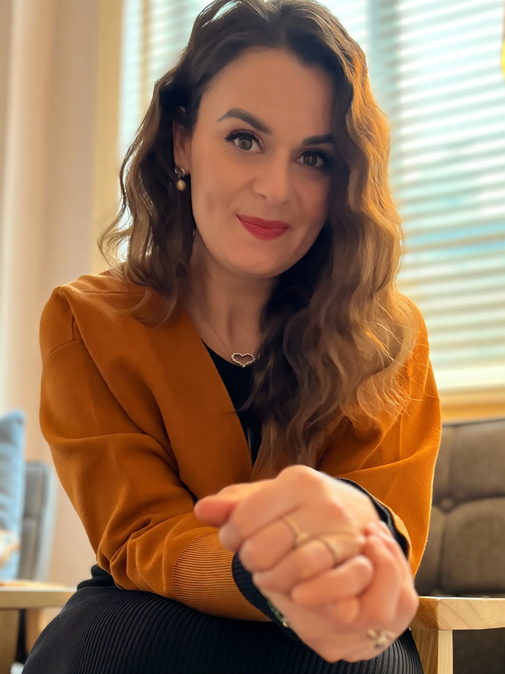 A woman therapist with wavy brown hair, red lipstick, and pearl earrings, wearing an orange blazer, is sitting indoors with her hands clasped in front of her, smiling at the camera. Mariana Aleksieva counselling