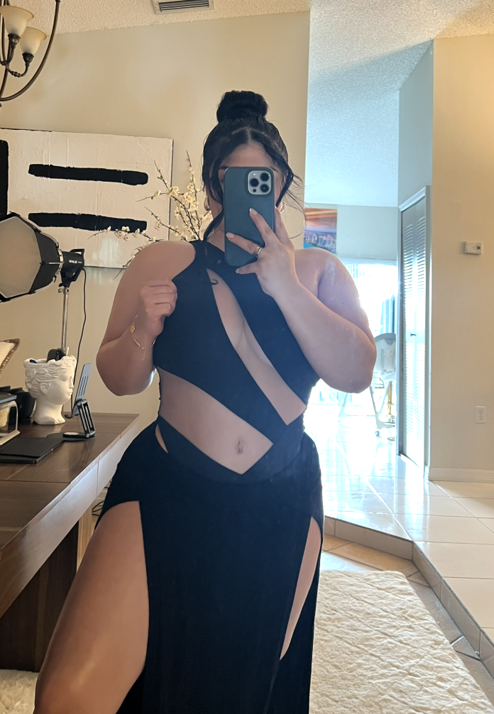Mujer con vestido negro con aberturas y tirantes, tomando selfie en espejo dentro de casa con varias decoraciones y muebles.