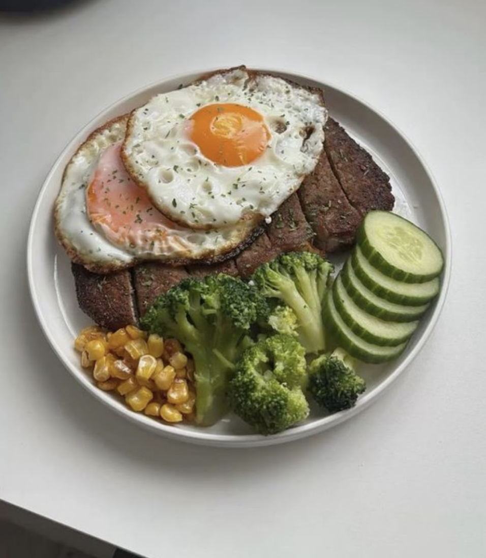 Plato con huevos fritos, carne asada, pepinos en rodajas, brócoli y maíz cocido.