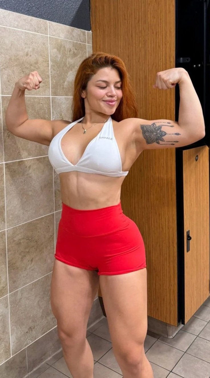 Mujer joven con cabello rizado y rubio, en ropa deportiva blanca y rojos, posando en un vestidor, mostrando sus músculos.»