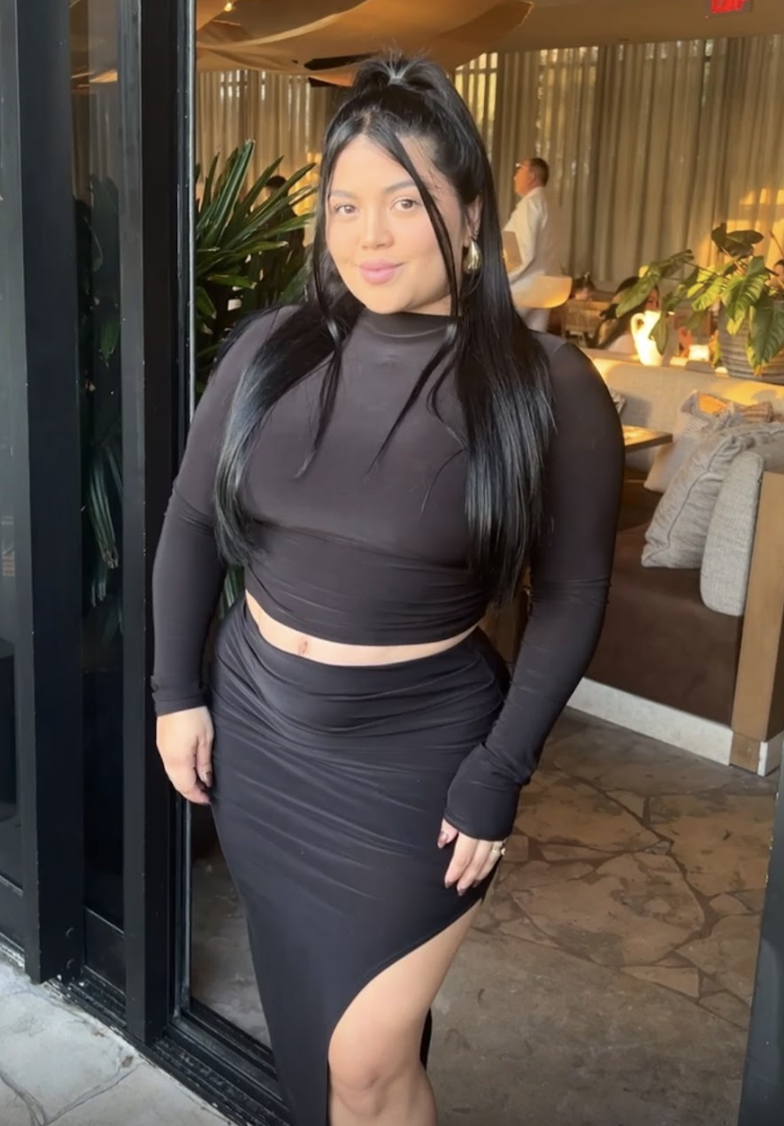 Mujer con vestido negro en un restaurante o cafetería, rodeada de plantas y decoraciones modernas.
