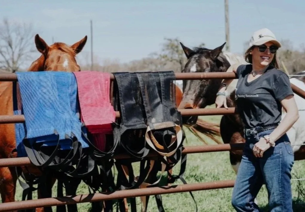 Woman wearing sunglasses and a hat standing next to horses at a farm, with colorful hammocks draped over a metal fence.