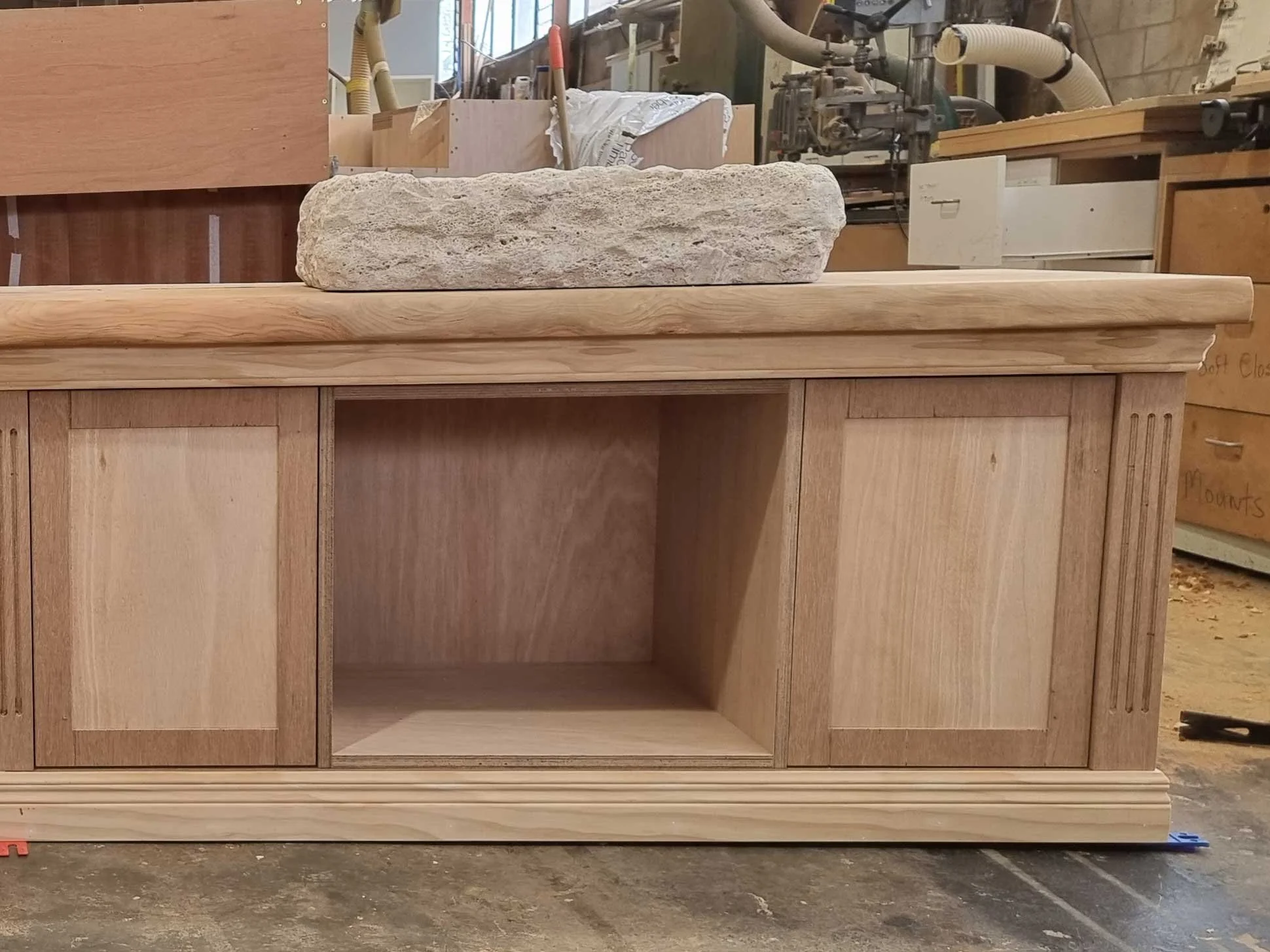 Wooden cabinet with two closed doors and an open space in the middle, topped with a large granite stone, in a woodworking shop with tools and equipment in the background.