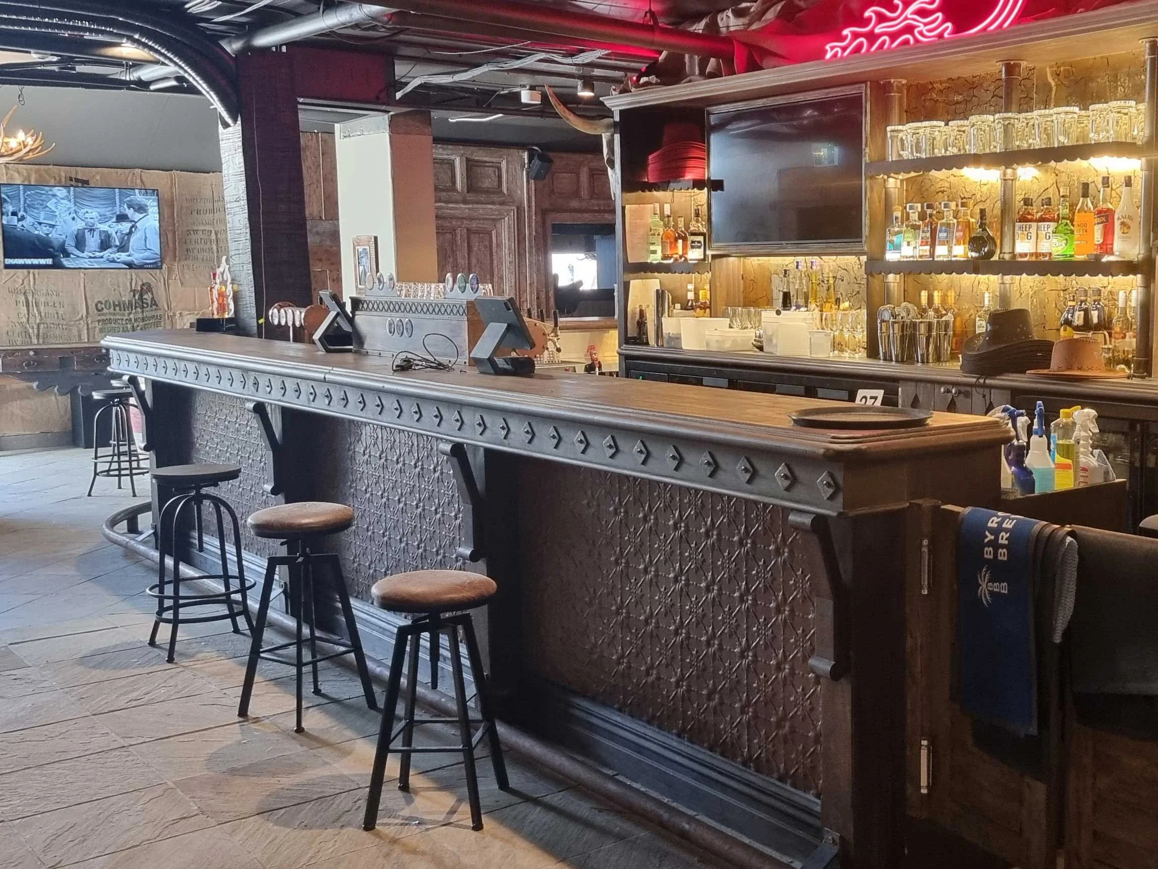 Empty bar counter with wooden bar, bar stools, shelves with liquor bottles, glasses, and bar equipment in a dimly lit restaurant or pub.