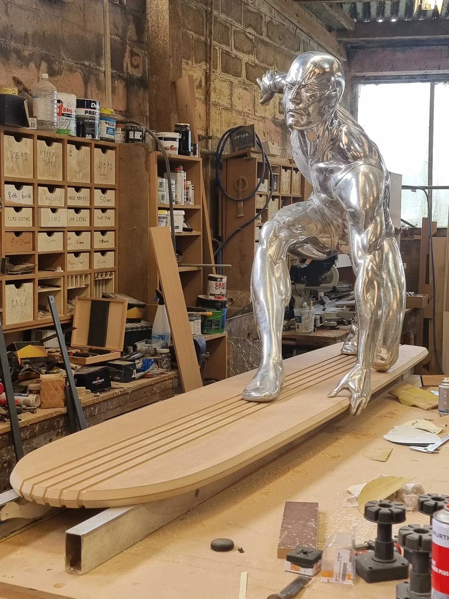 A sculpture of a muscular man in a crouching pose, made from a shiny material, positioned on a wooden surfboard in a woodworking workshop.